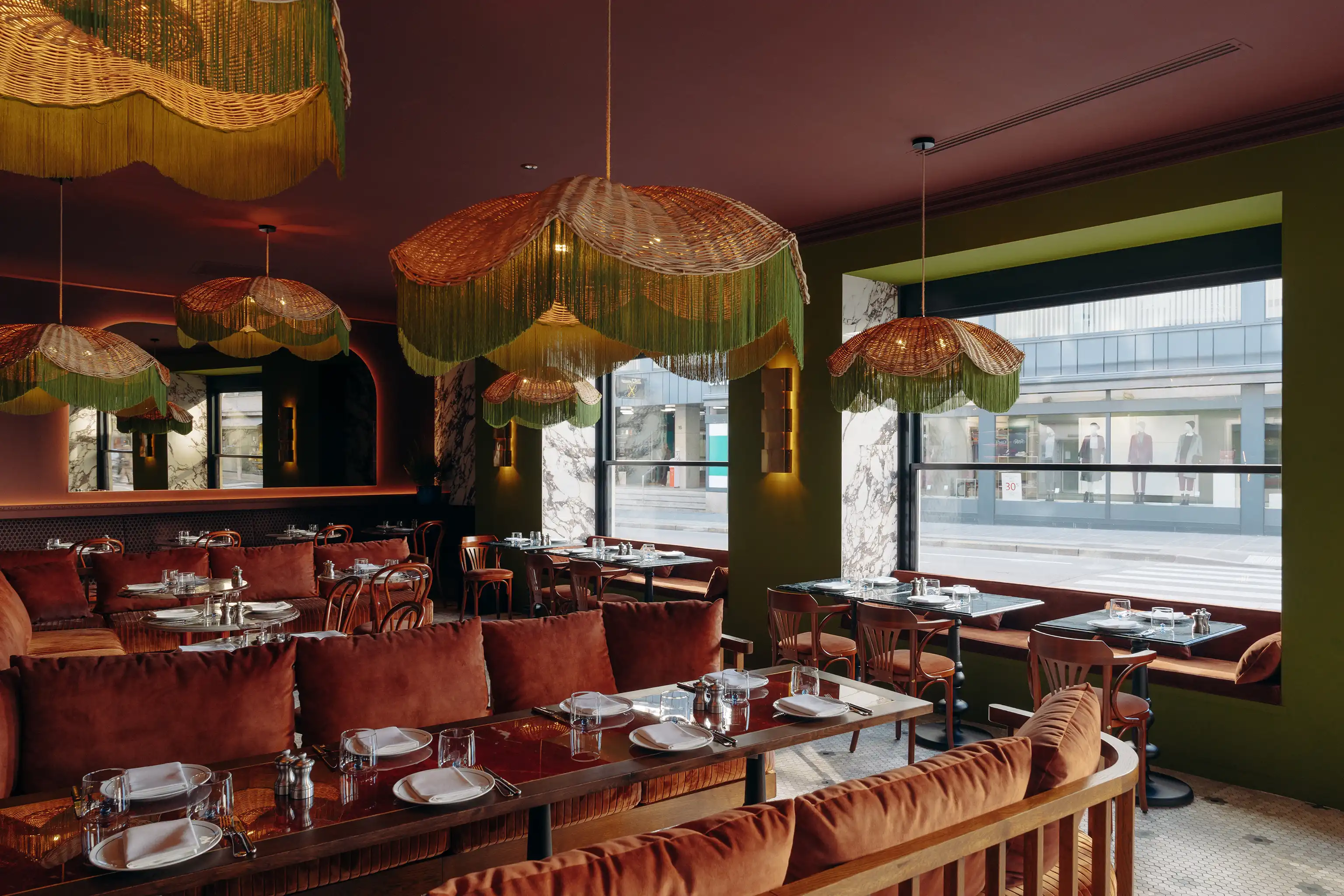 Cozy restaurant interior with velvet seating, wooden chairs, dark green walls, marble window frames, and decorative hanging lamps with green fringes.