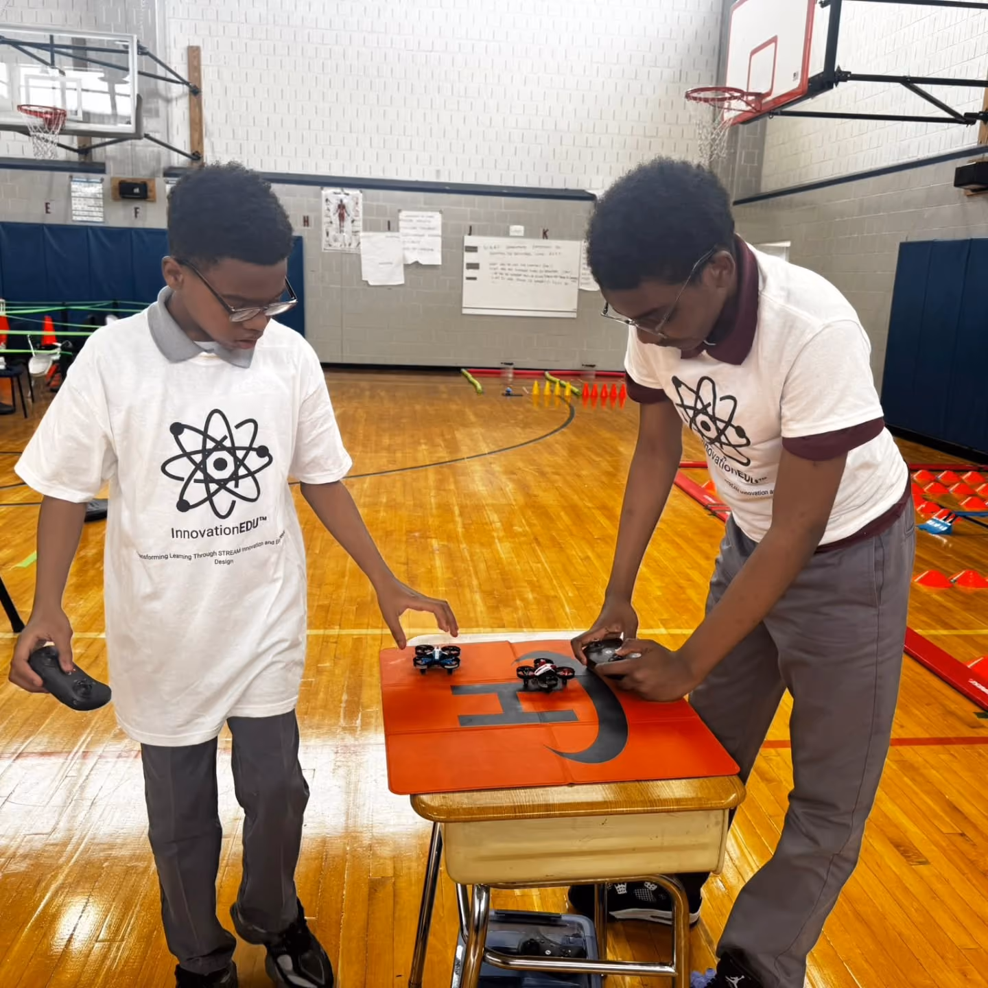 Two students operating mini drones on a landing pad in gym