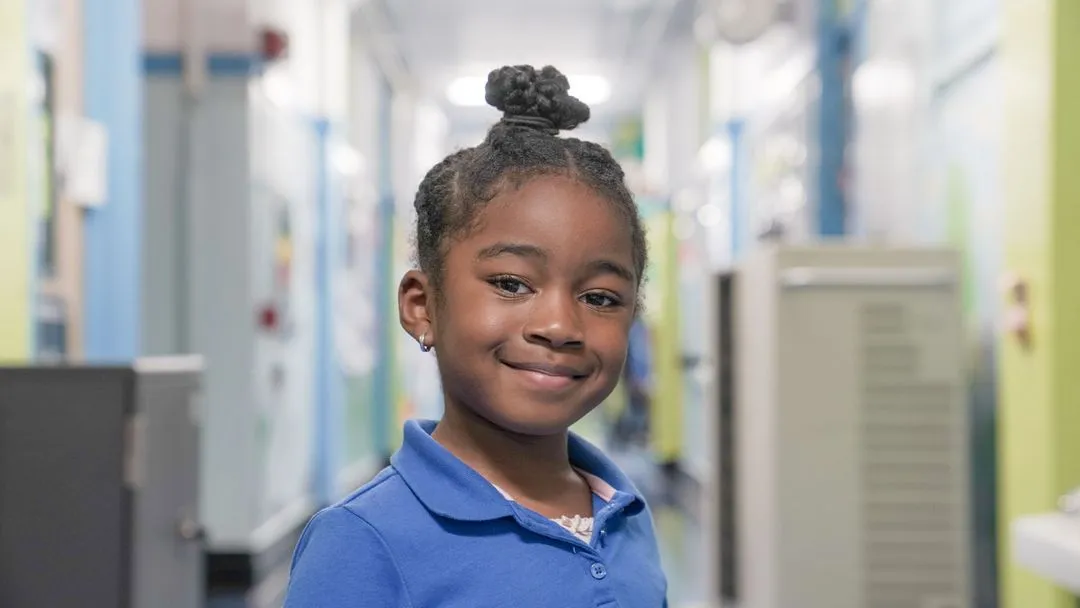 A smiling girl wearing a blue polo shirt stands in a bright school hallway, facing the camera.