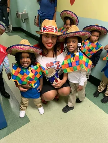  A group of young children and an adult dressed in colorful costumes for a cultural event. The children wear straw hats and vibrant square-patterned ponchos, and the adult kneels in the center holding maracas.








