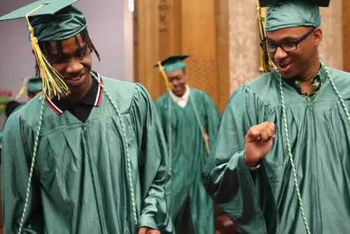 – Two students in green caps and gowns smiling and talking during a graduation ceremony.