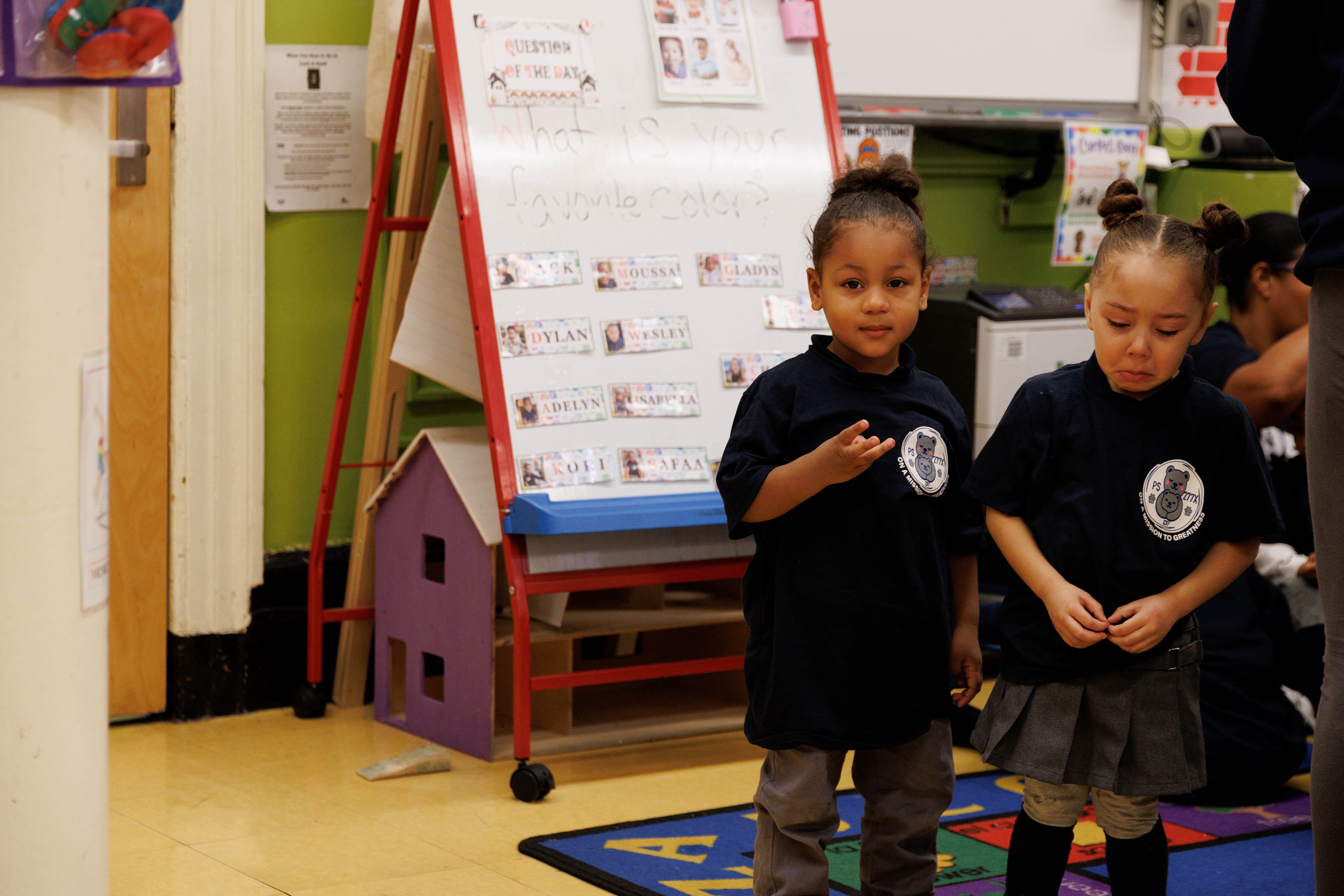 Two young children in matching navy shirts standing on a colorful alphabet rug in a classroom with a whiteboard behind them.