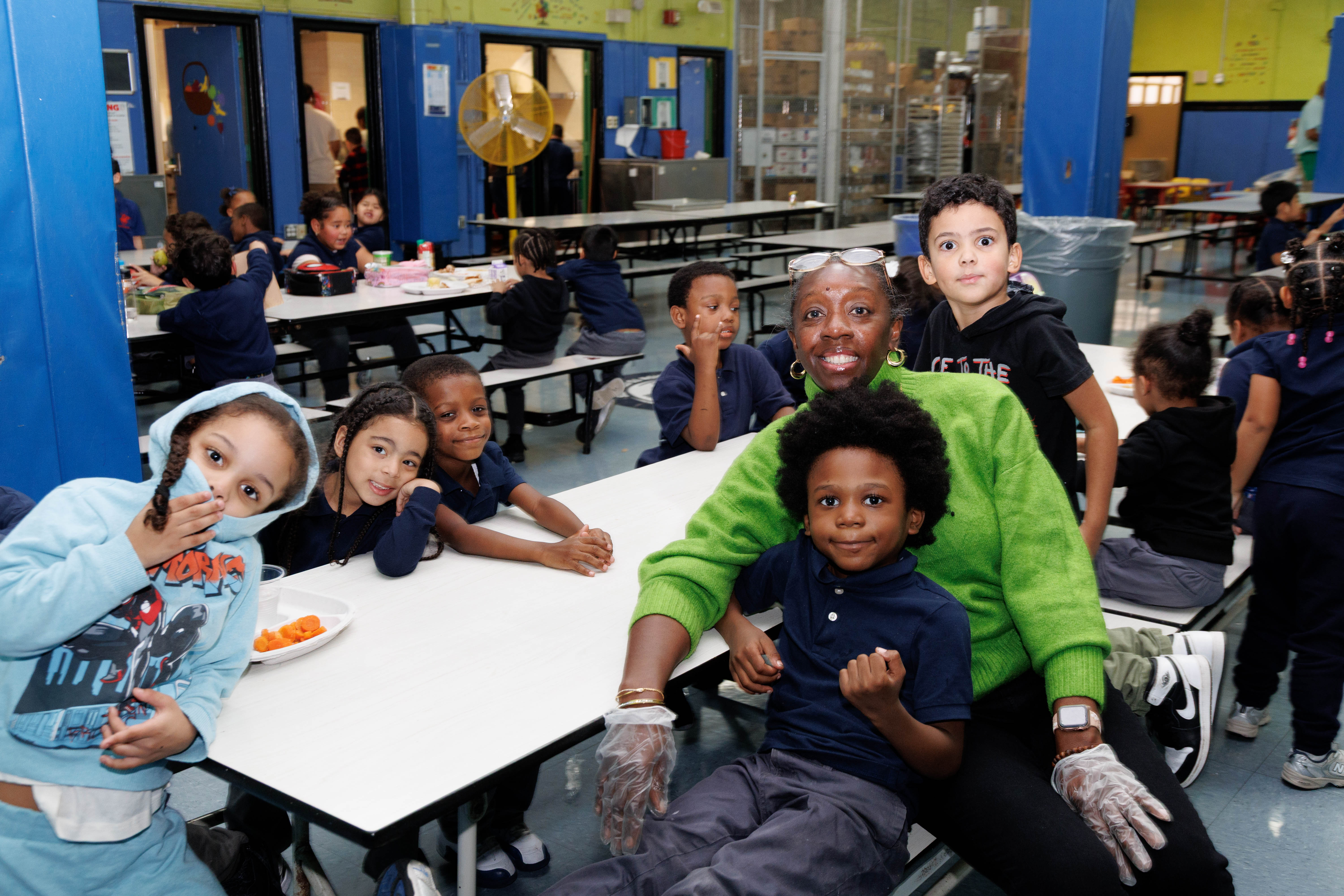 Group of children and a smiling adult in a green sweater wearing plastic gloves sitting at tables in a brightly colored cafeteria.