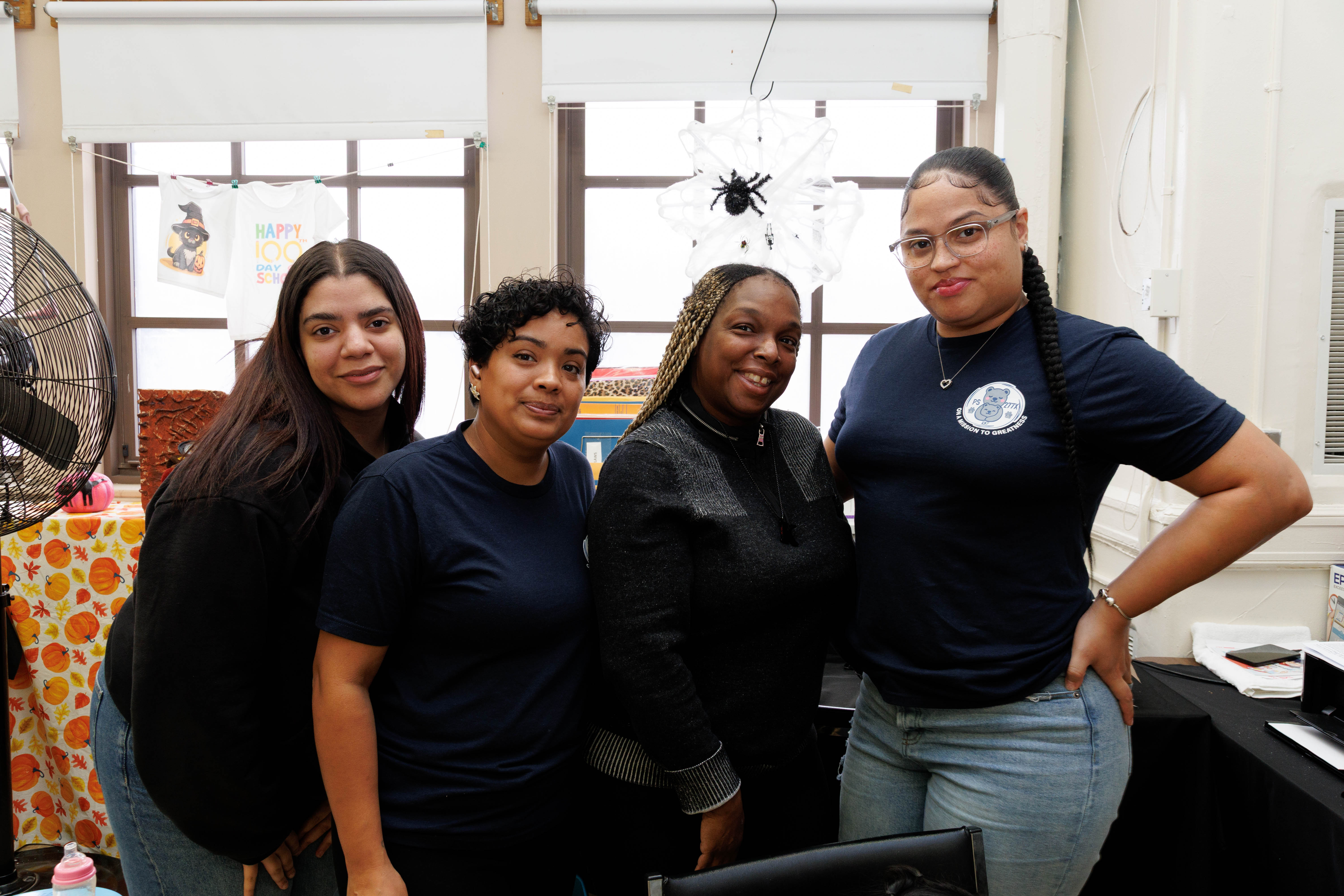 Four women standing closely together, smiling in a room decorated with a spider web and Halloween-themed items.