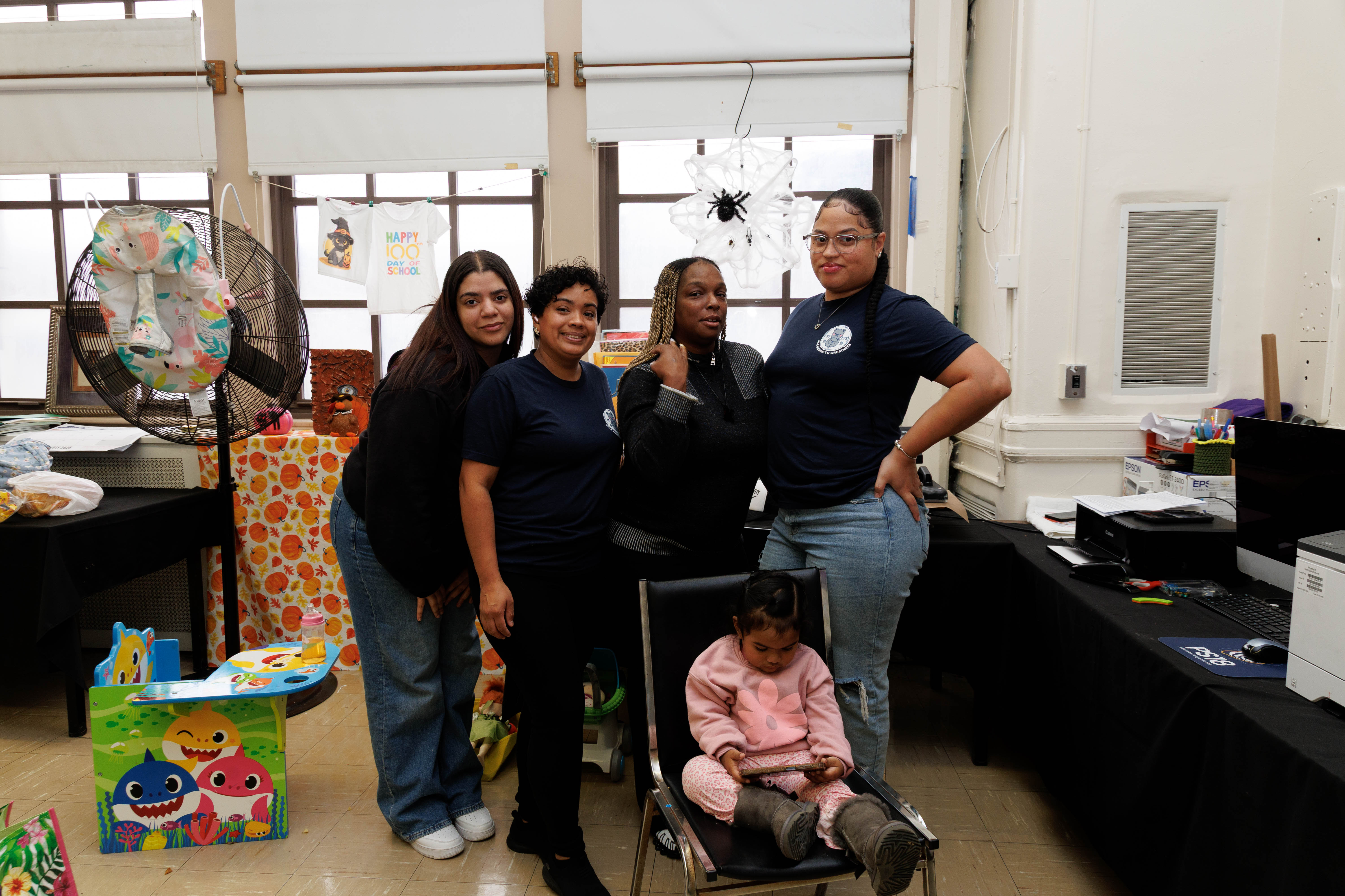 Four women standing behind a chair where a young girl in a pink sweater and boots is sitting and looking at a tablet in a room decorated with colorful children's items.