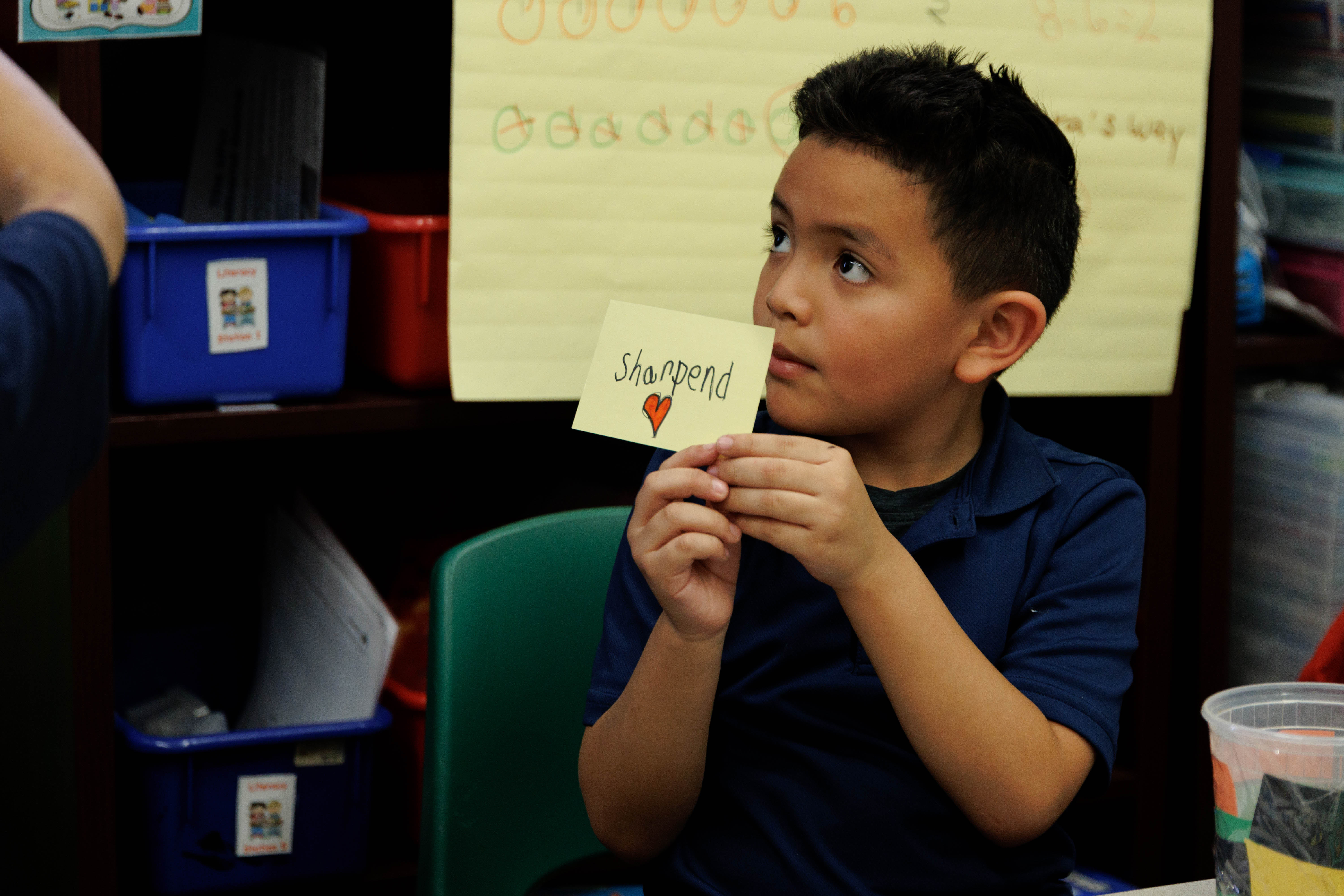 Young boy sitting in a classroom holding a sticky note with the word 'Sharpend' and a red heart drawn below it.