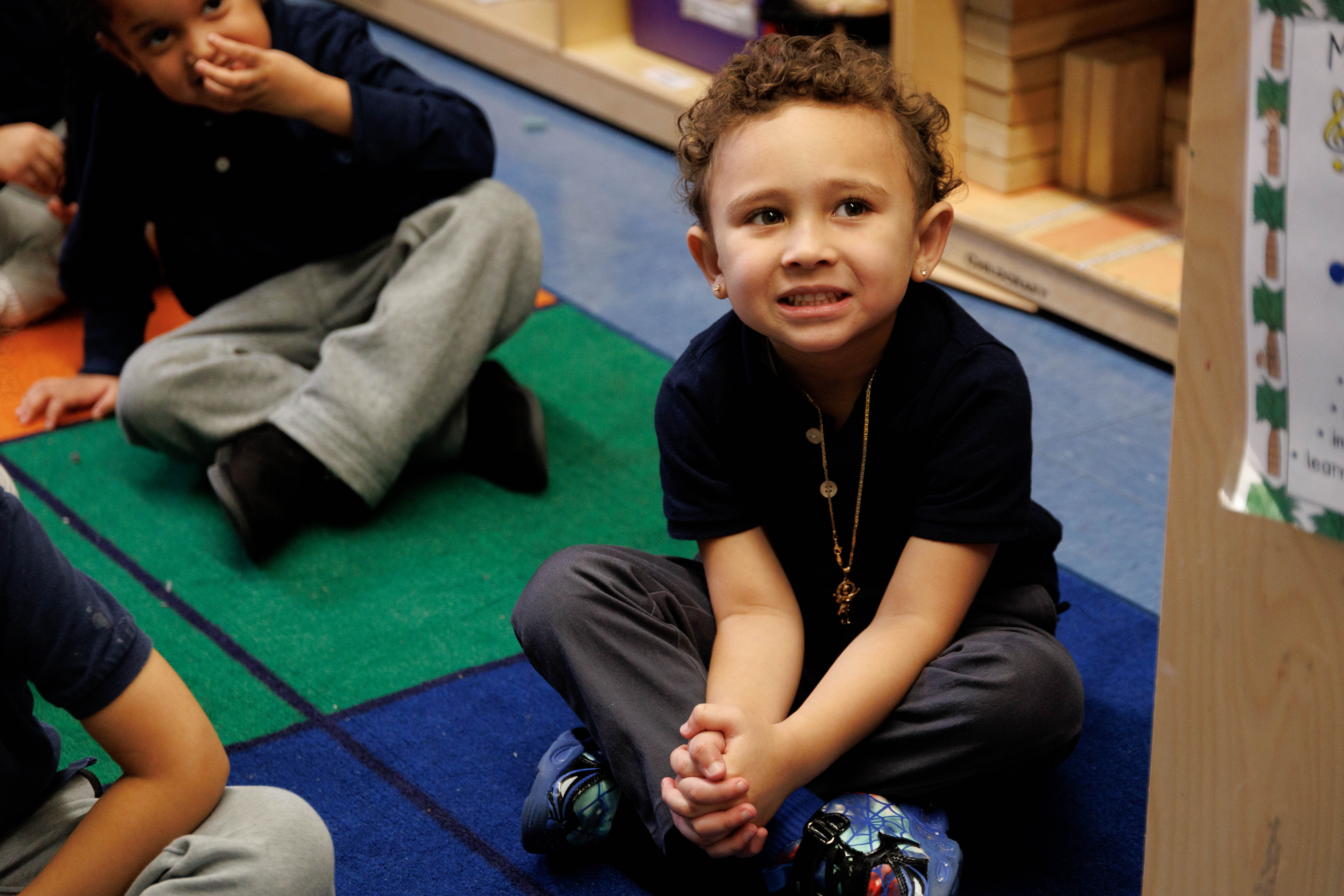 Young child sitting cross-legged on a colorful classroom rug with hands clasped, looking slightly nervous.