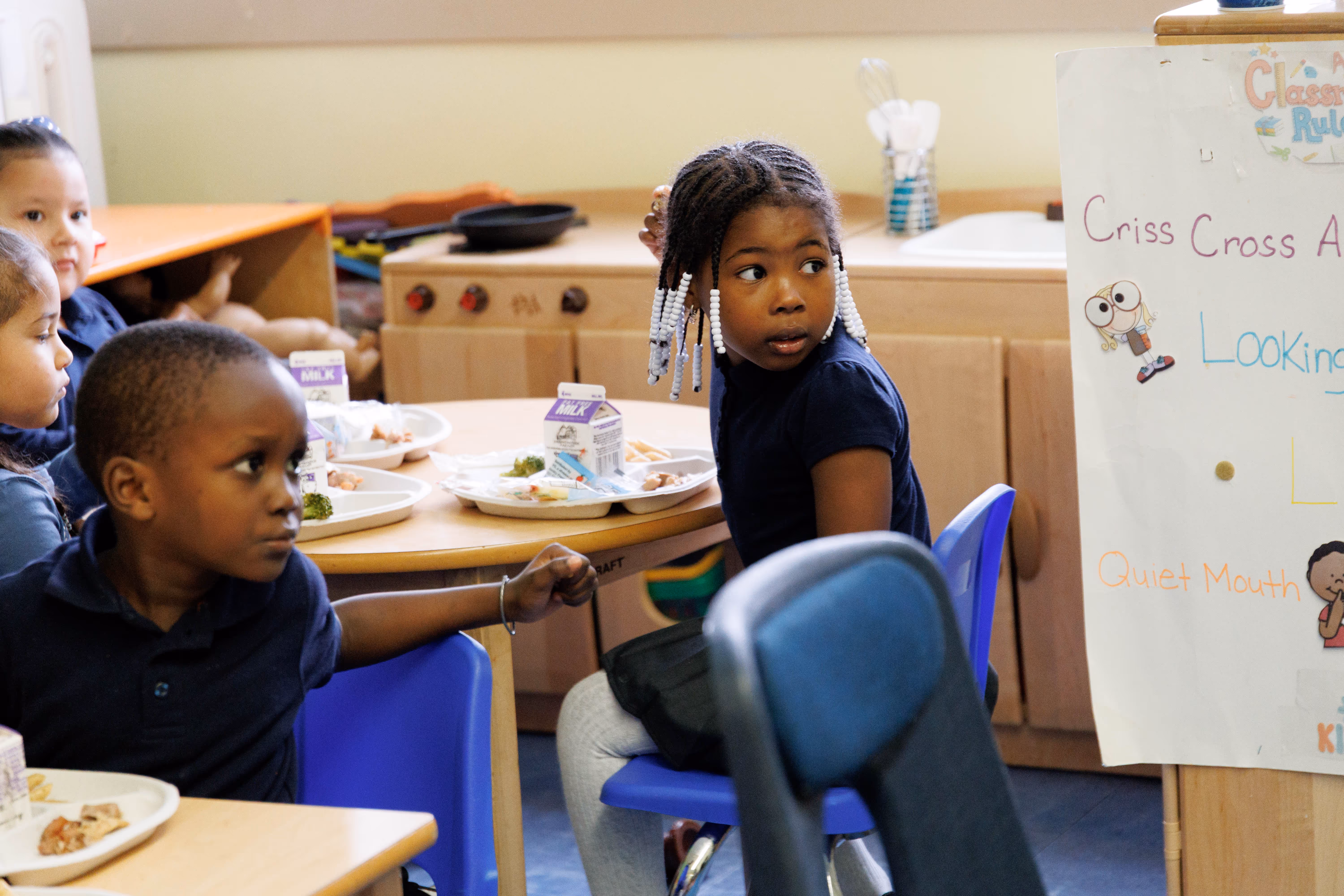 Young children sitting at tables having a meal with milk cartons in a classroom setting.