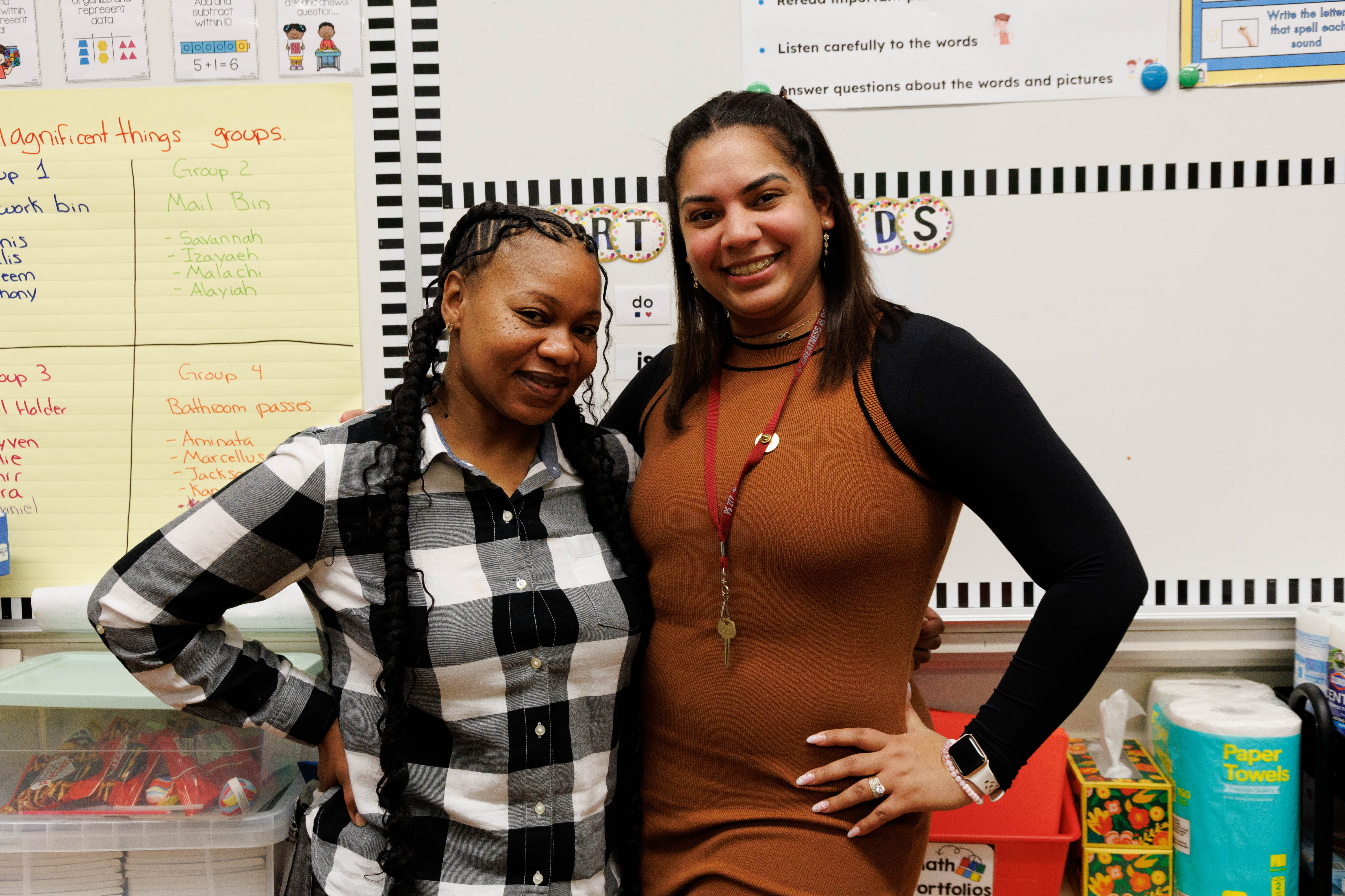 Two women smiling and posing together in a classroom with educational posters and supplies in the background.