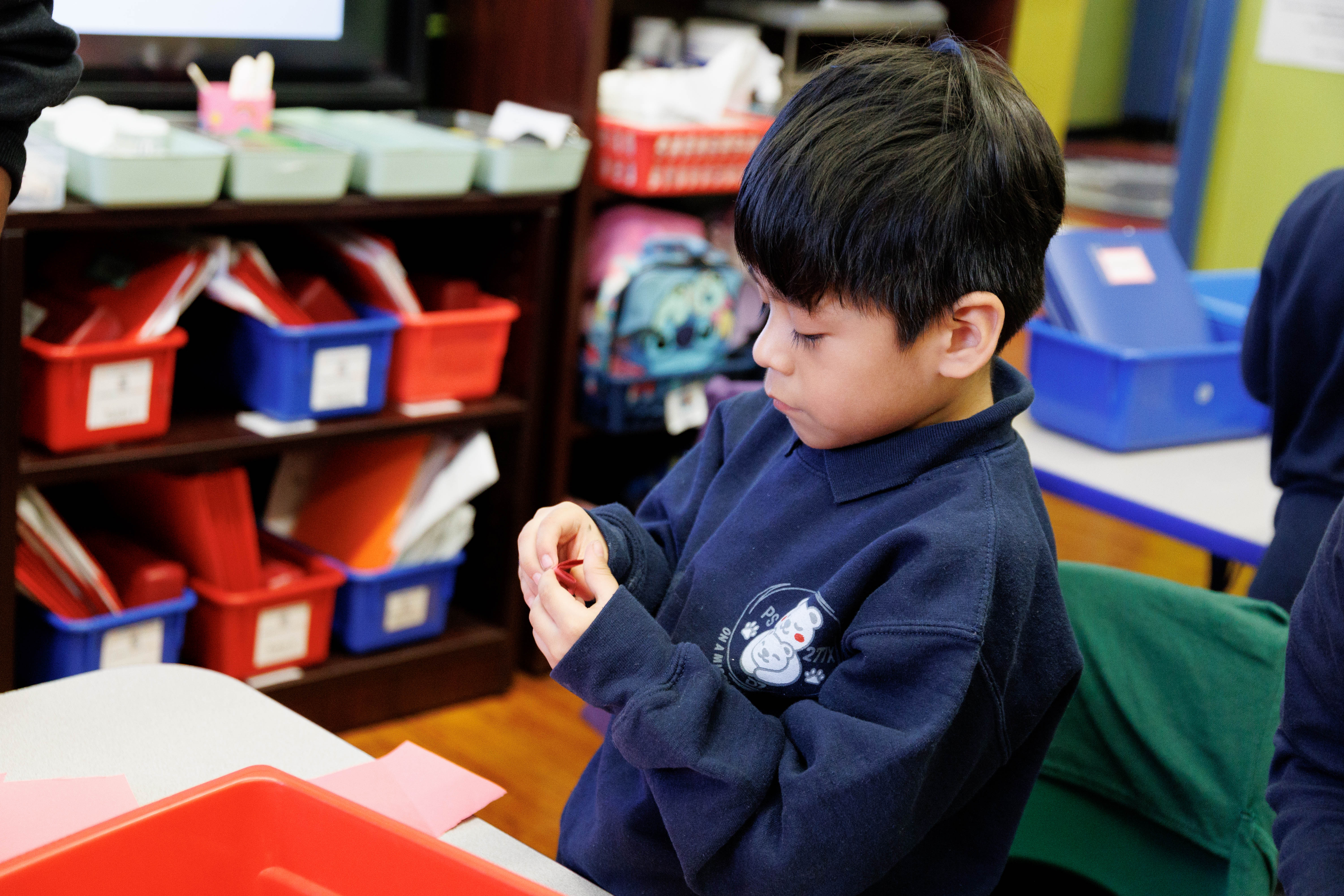 Young boy in navy blue sweatshirt focused on folding red paper at a classroom table.
