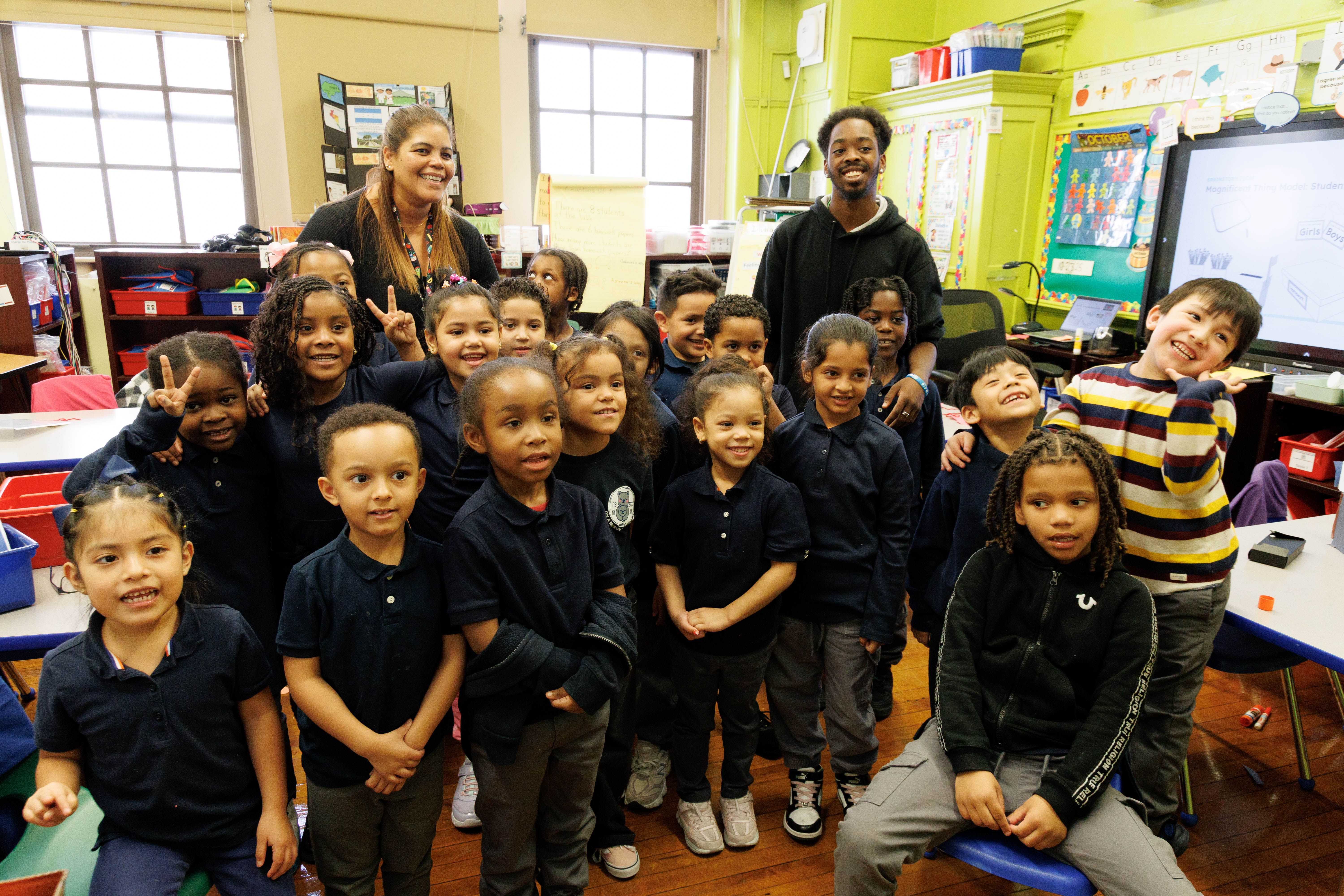 A group of diverse young children and two teachers smiling together in a colorful classroom.
