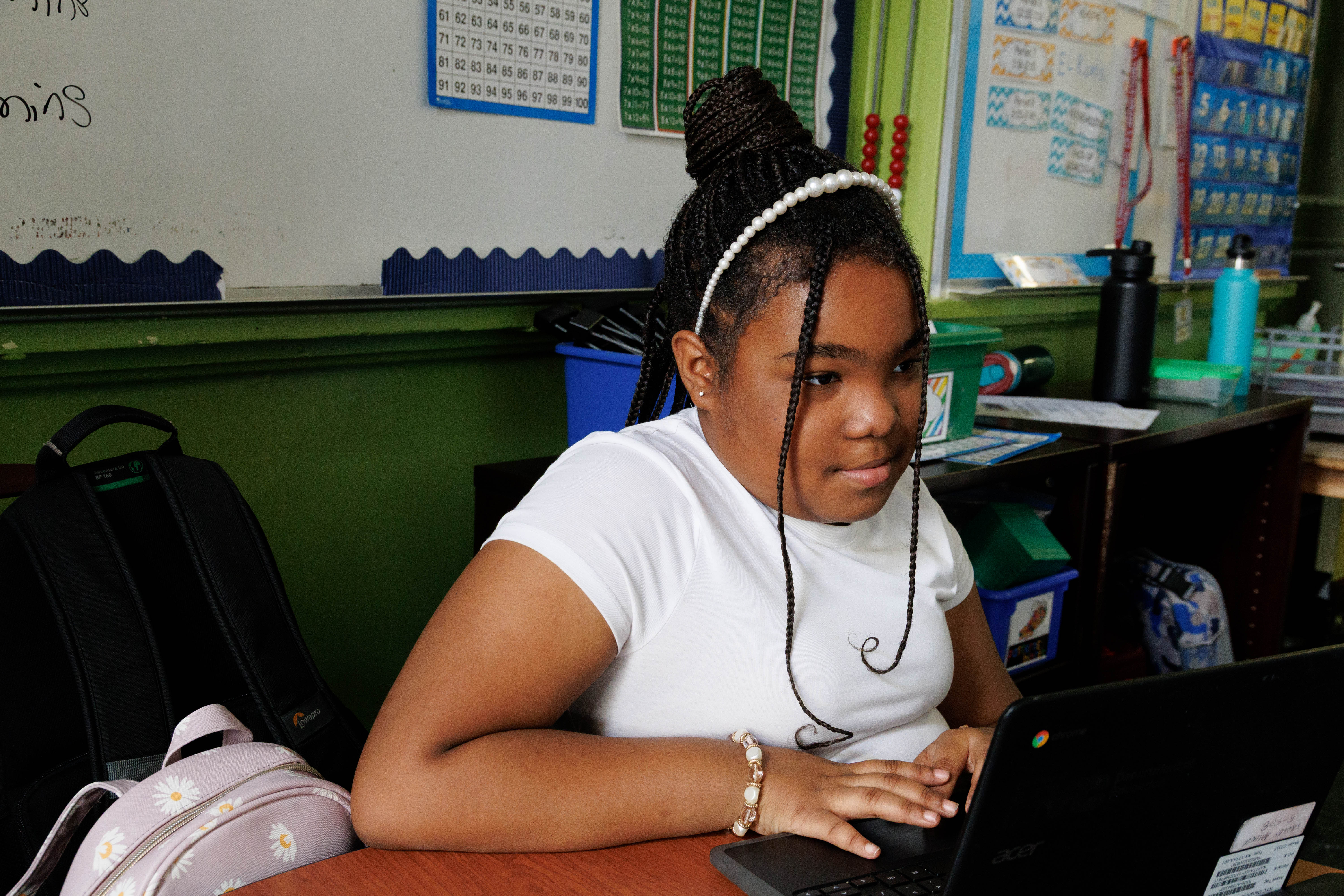 Young girl with braid and pearl headband using a laptop in a classroom.