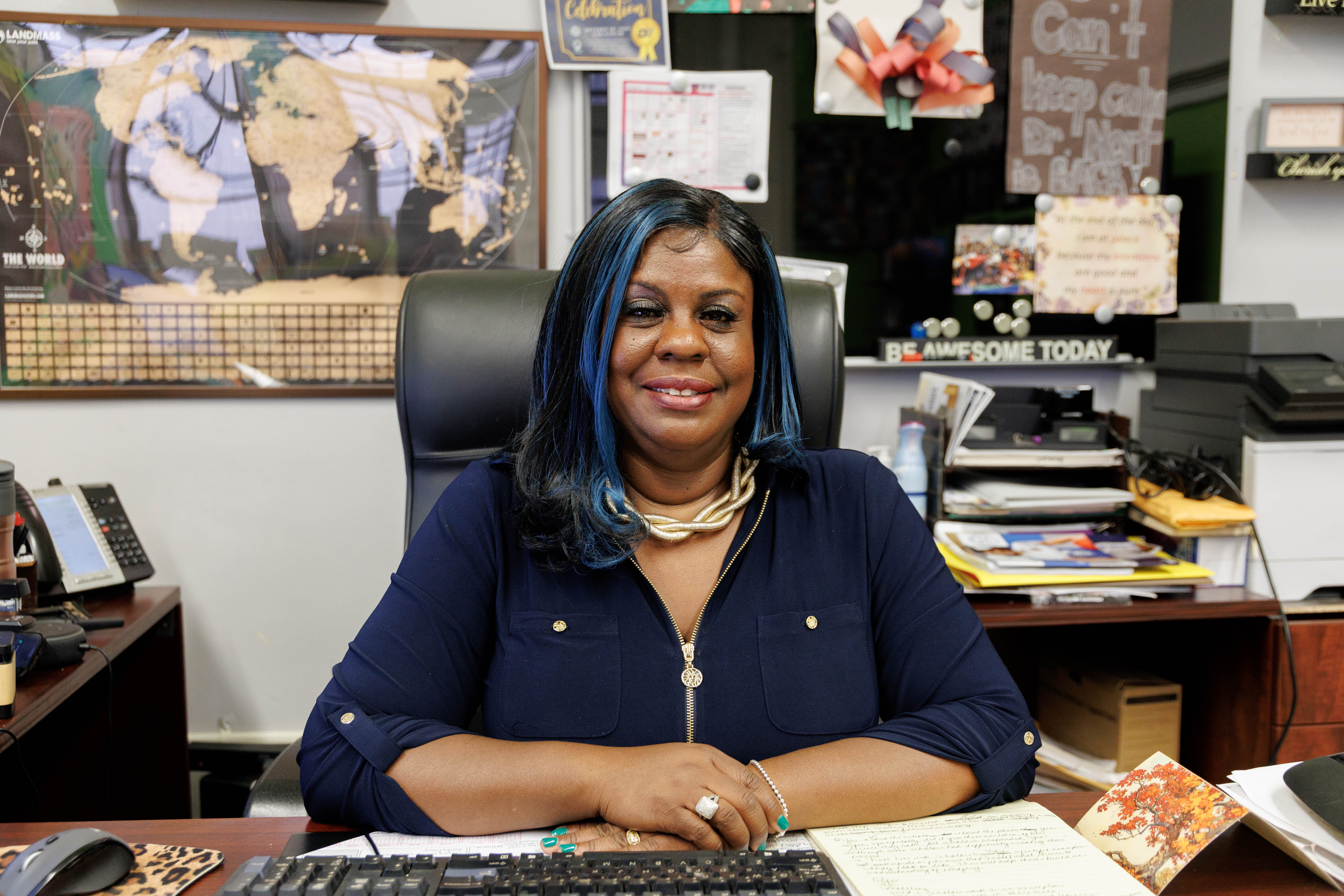 Smiling woman with blue-streaked hair sitting at an office desk with a keyboard, notebook, and map on the wall behind her.