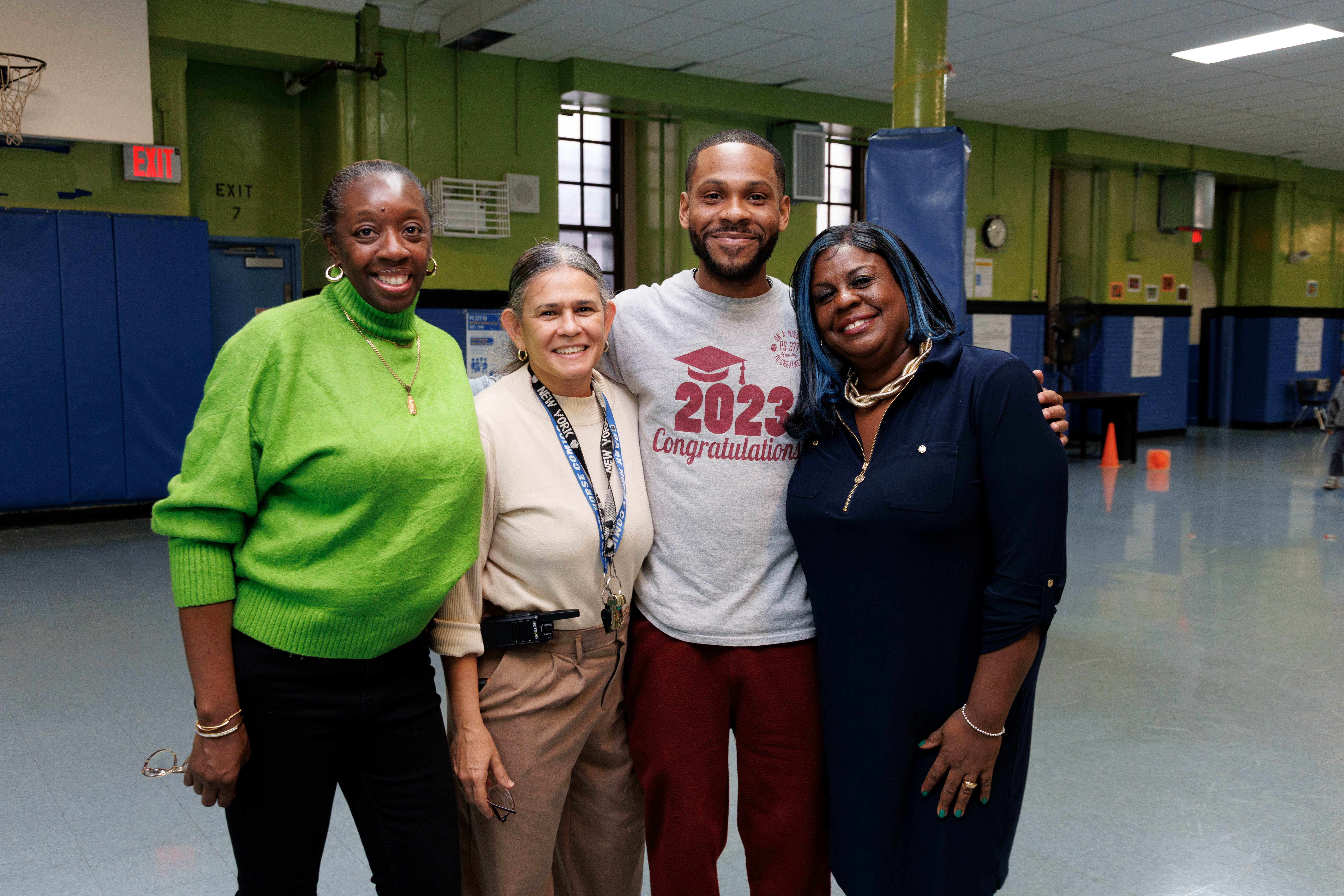Four diverse adults smiling and standing closely together in a gymnasium, with one wearing a gray 2023 congratulations T-shirt.