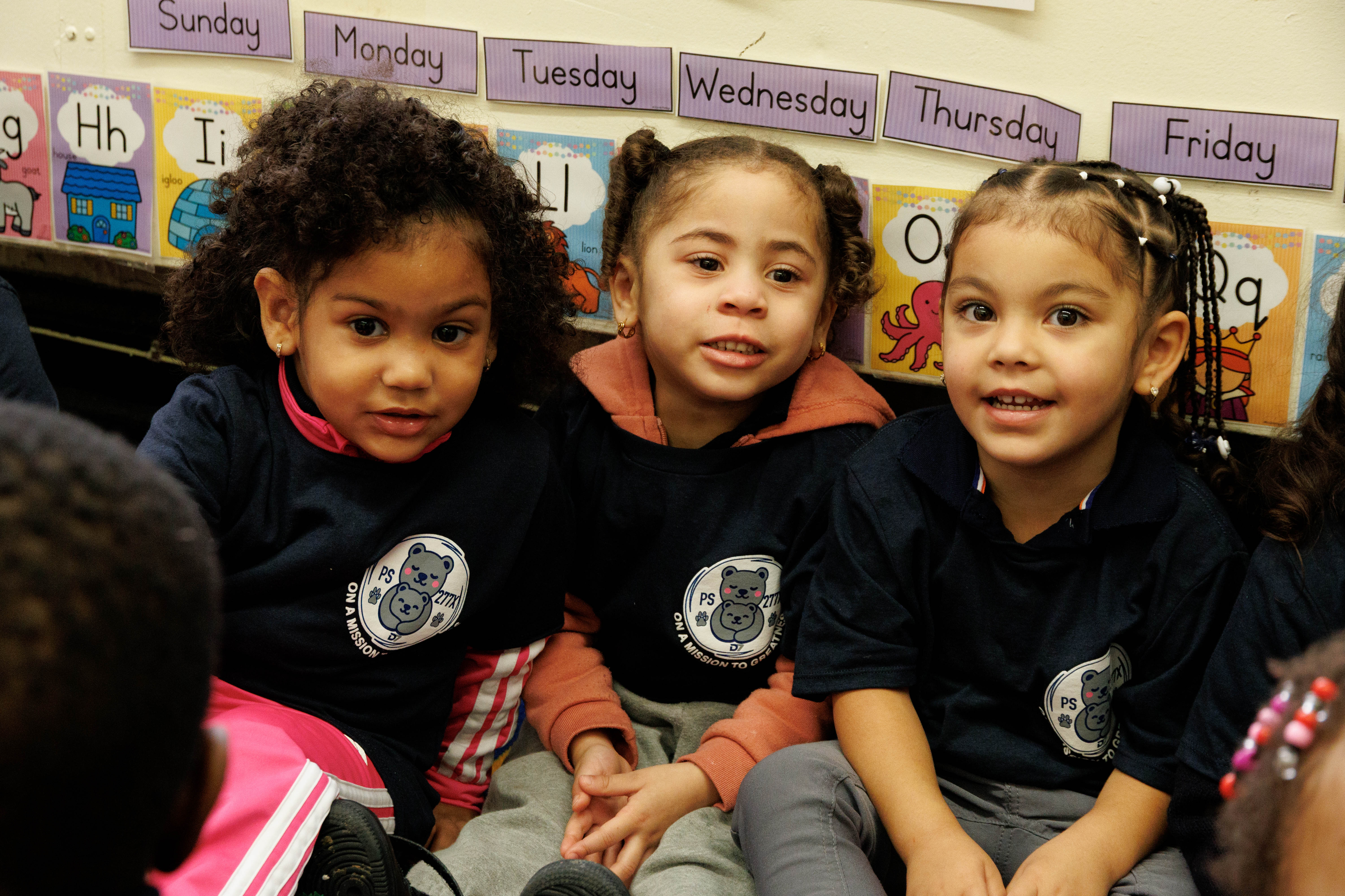 Three young children sitting closely together in a classroom with weekday labels on the wall behind them.