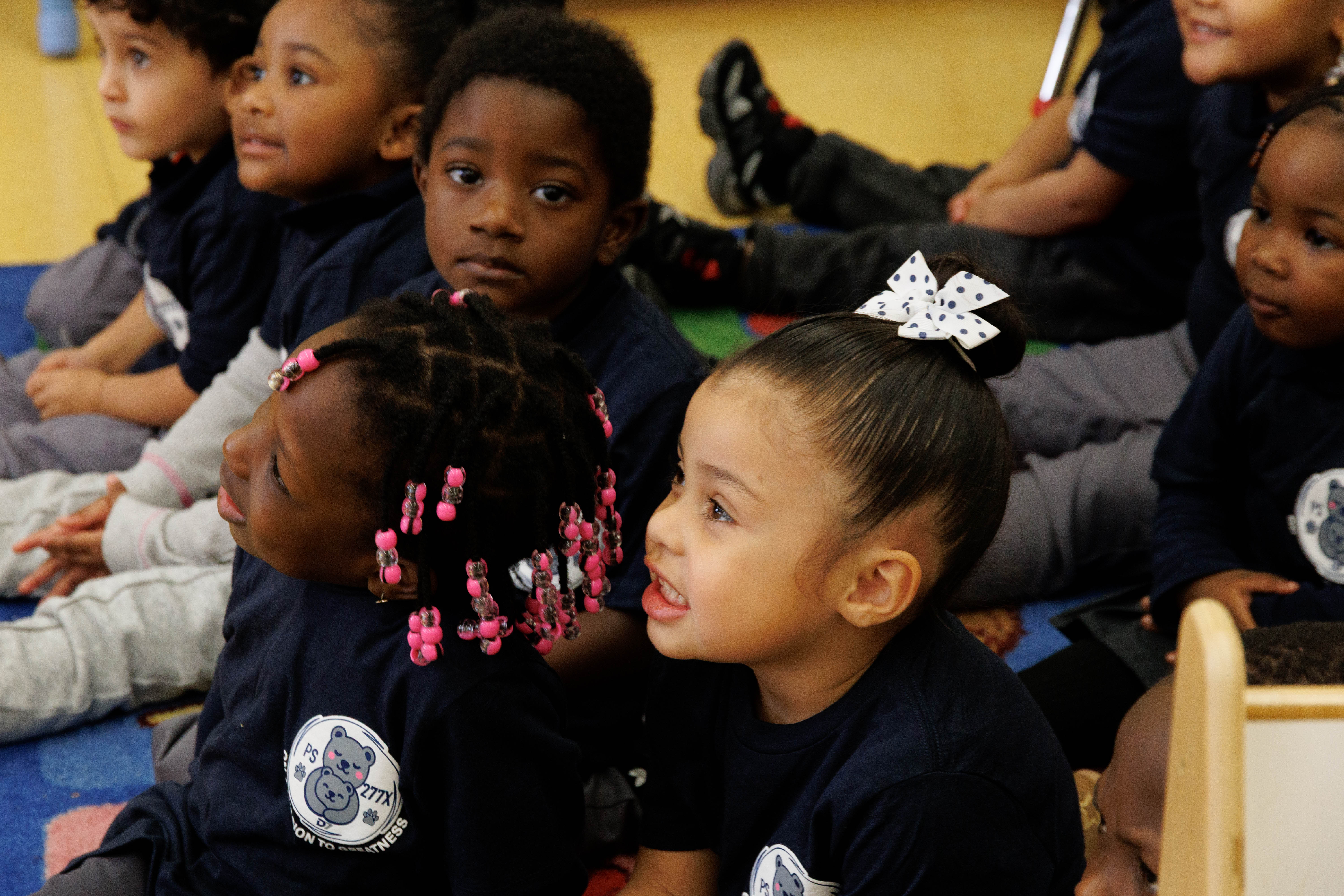 Group of young children in matching navy shirts sitting attentively on a classroom carpet.