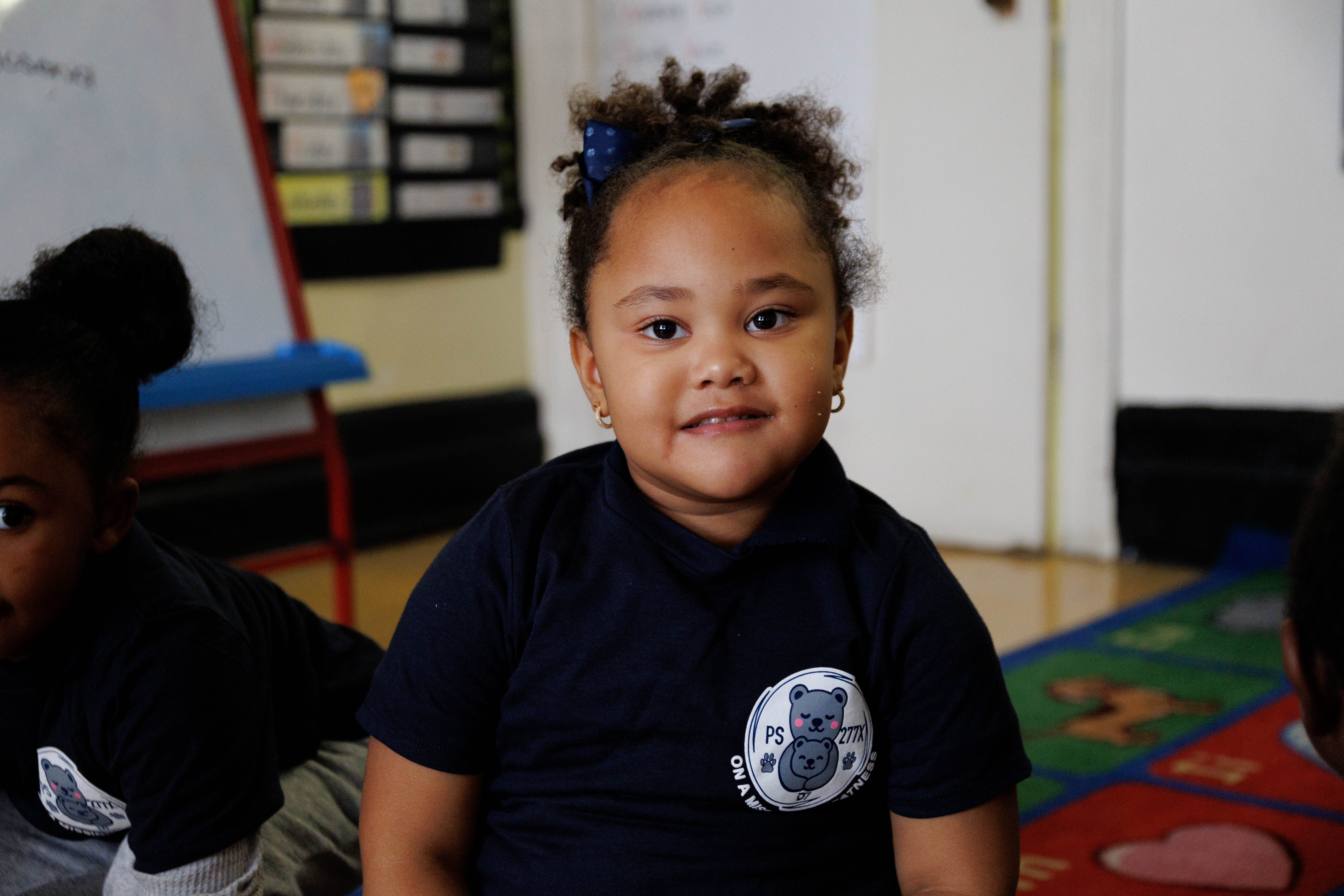 Young girl with curly hair and a blue bow wearing a navy shirt with a bear logo, sitting in a classroom.