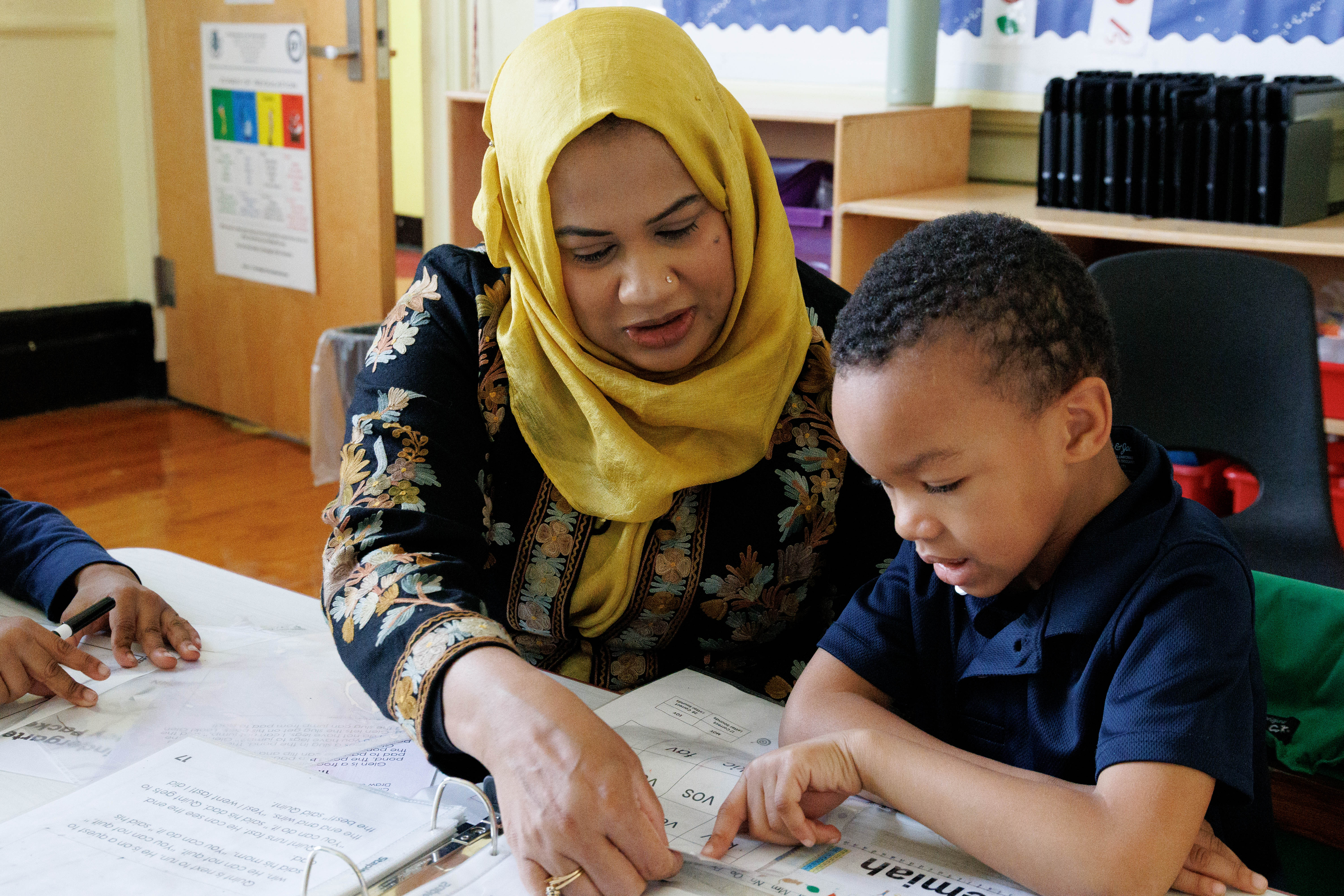 Teacher in yellow hijab guiding young student through reading worksheet