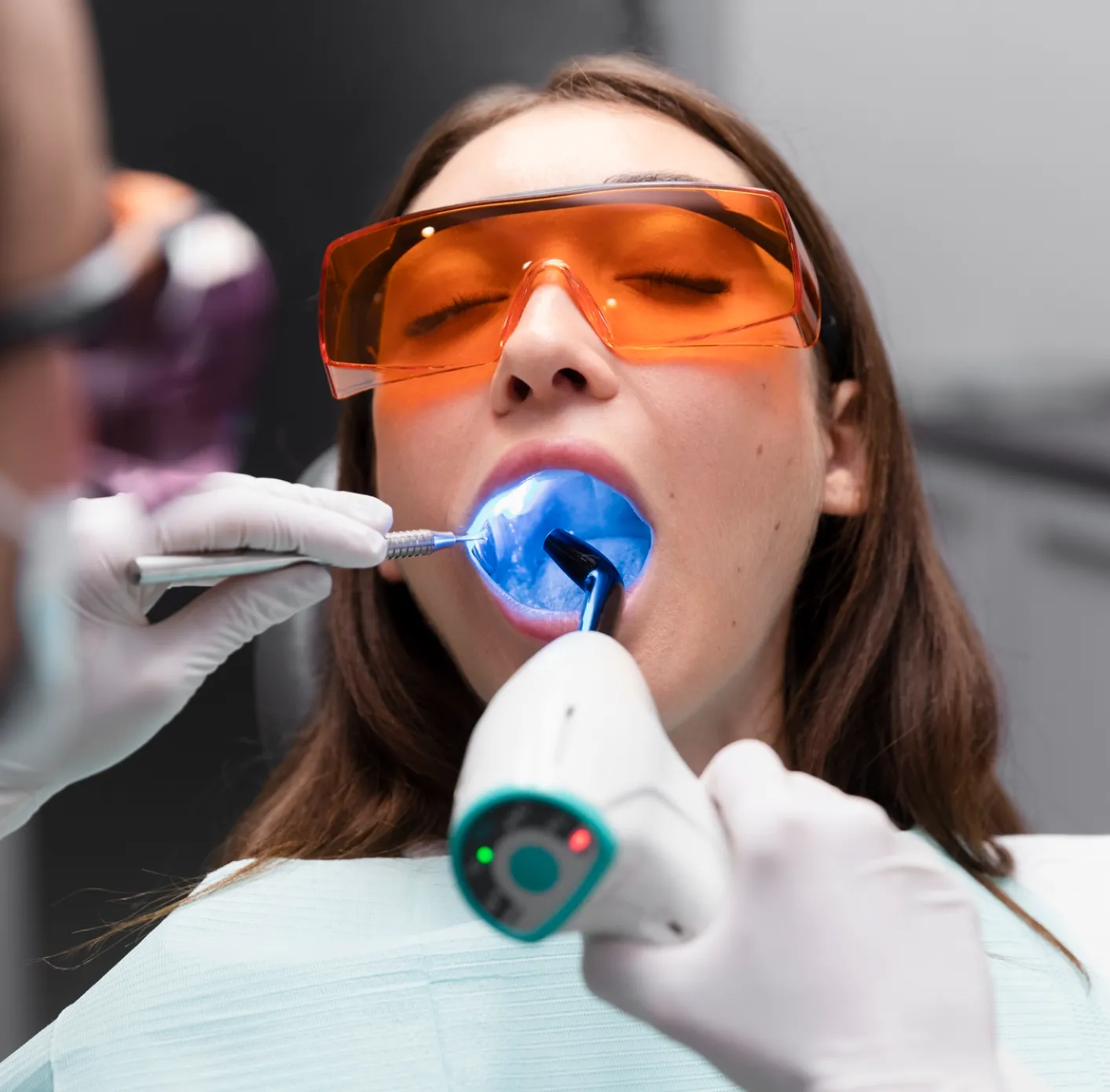 Patient wearing orange protective glasses undergoing dental treatment with blue curing light and dental tool.