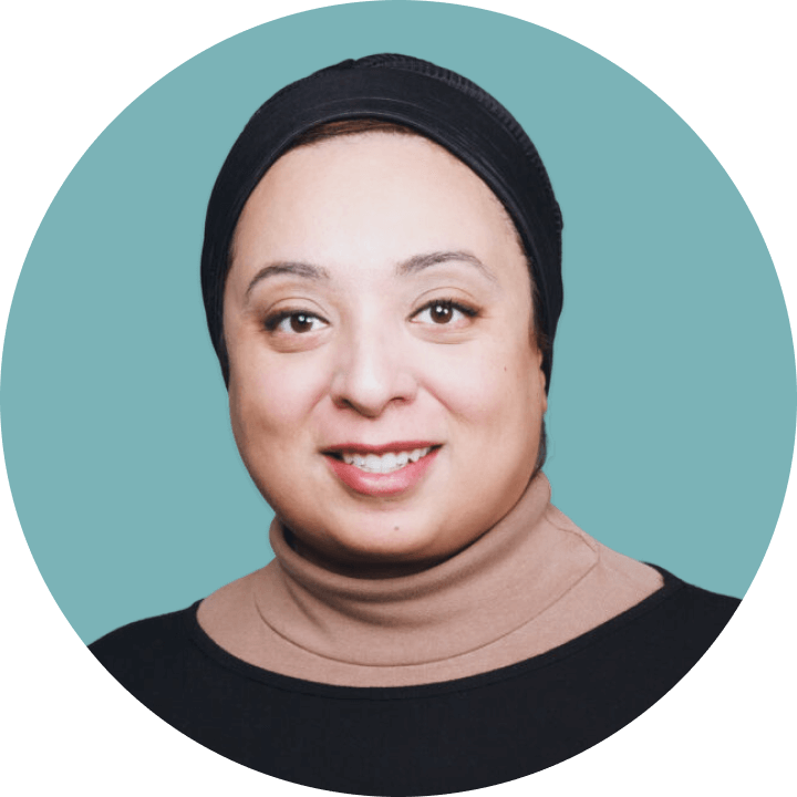 Portrait of a woman wearing a black headscarf and beige turtleneck, smiling against a blue background.