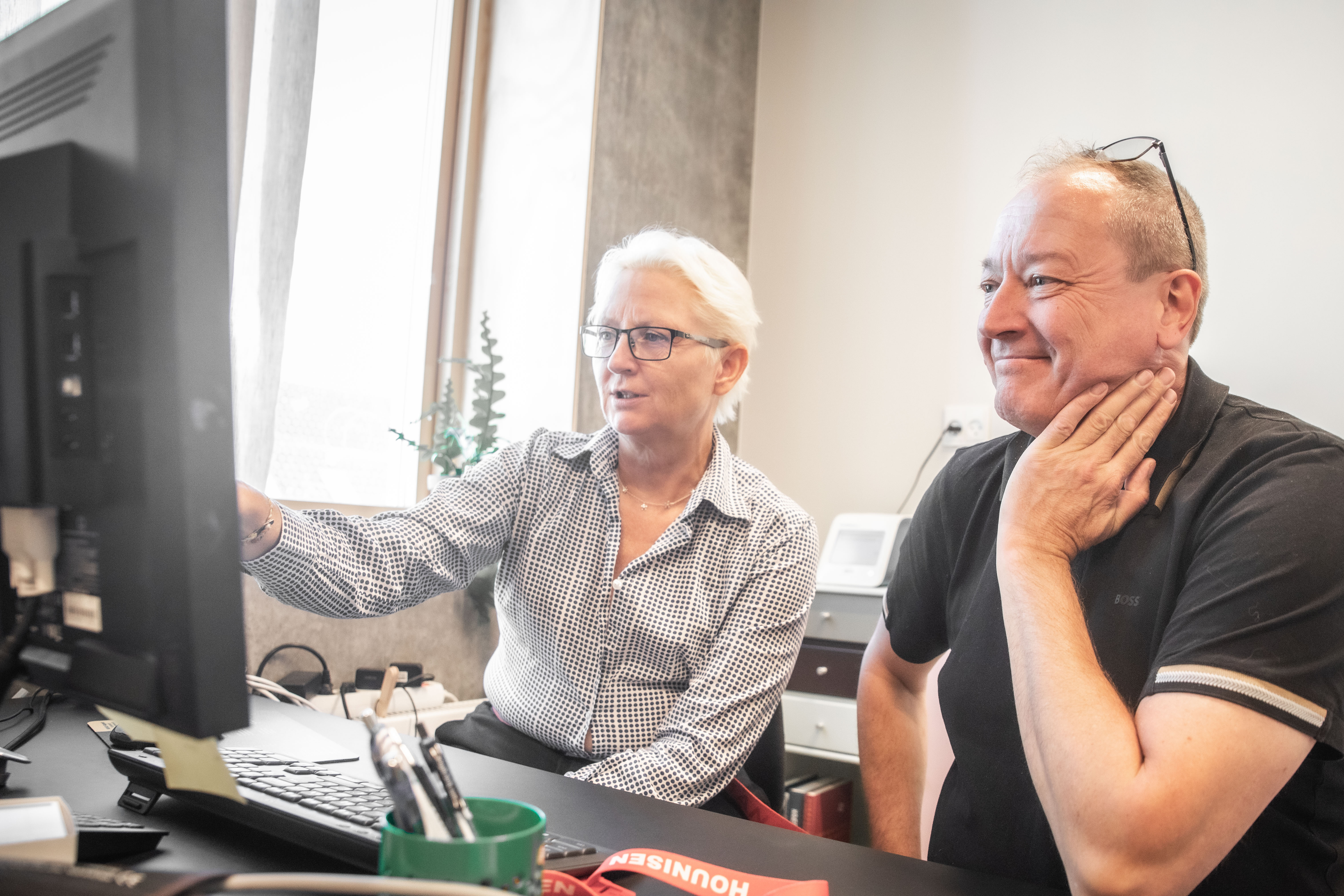 En kvinde med hvidt hår peger på en computerskærm, mens en mand i sort t-shirt lytter opmærksomt i et kontormiljø.