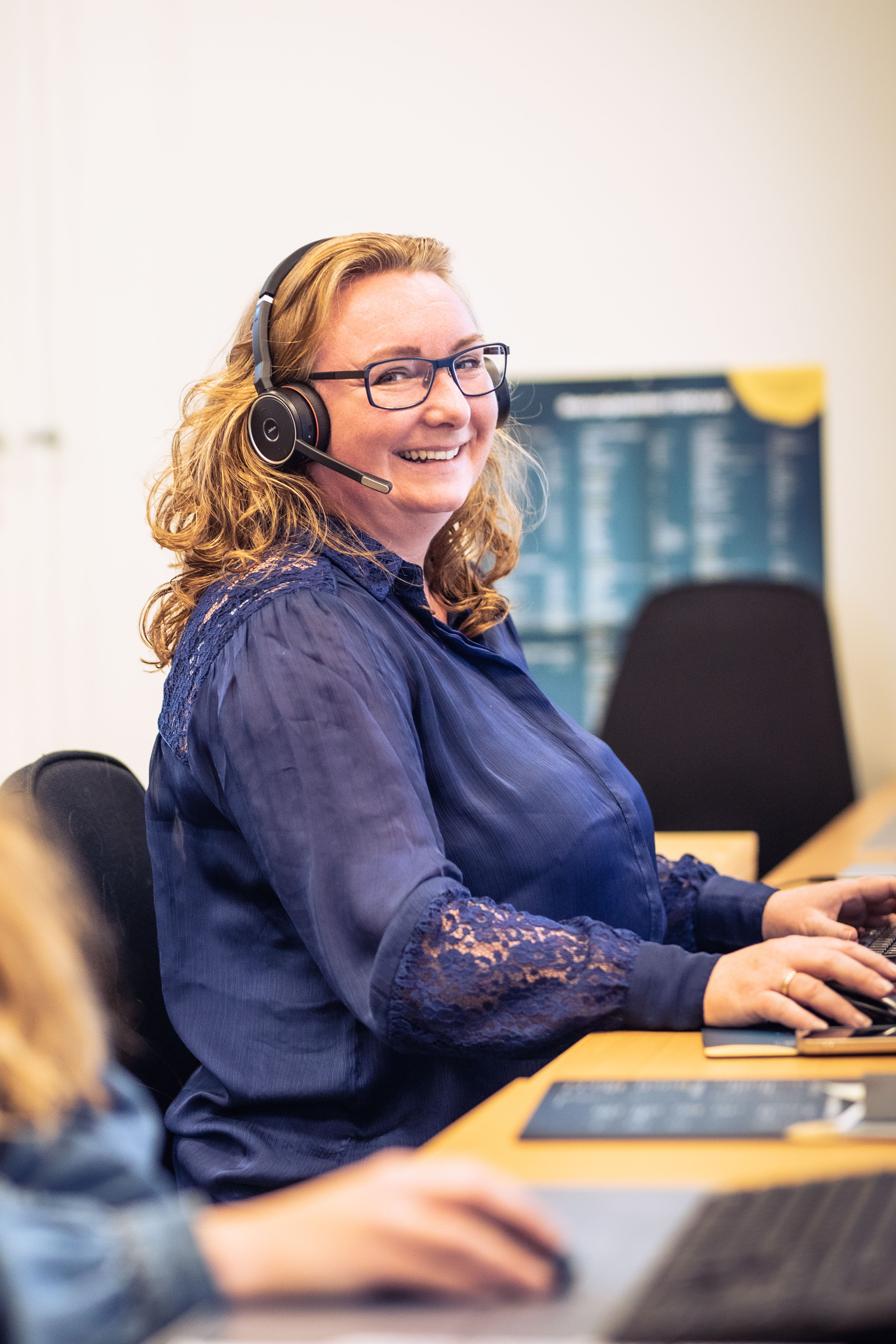 Smilende kvinde med headset og briller, der sidder ved en computer og arbejder i et kontormiljø.