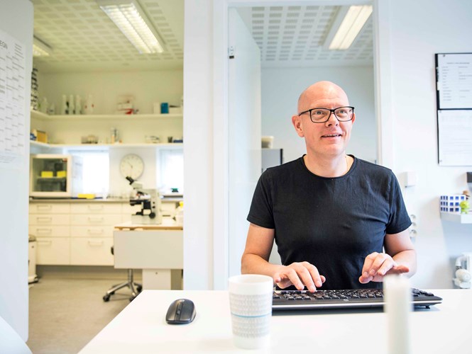 Mand med briller sidder ved et skrivebord og arbejder på en computer, i et lyst kontormiljø med laboratorieudstyr i baggrunden.