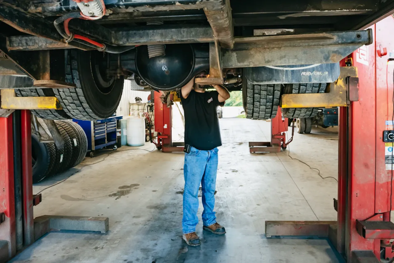 Technician performs preventive maintenance under a lifted truck, inspecting suspension and drivetrain components.