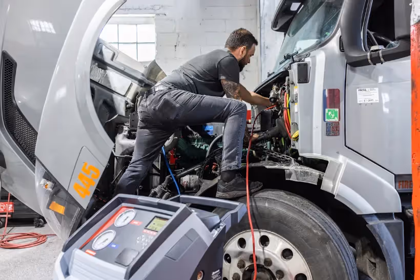 Technician performing HVAC service on semi-truck engine with cab tilted, diagnostic machine and “445” visible on hood.