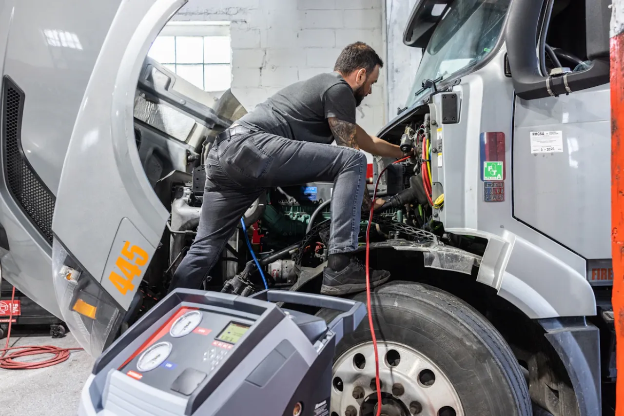 Technician performing HVAC service on semi-truck engine with cab tilted, diagnostic machine and “445” visible on hood.