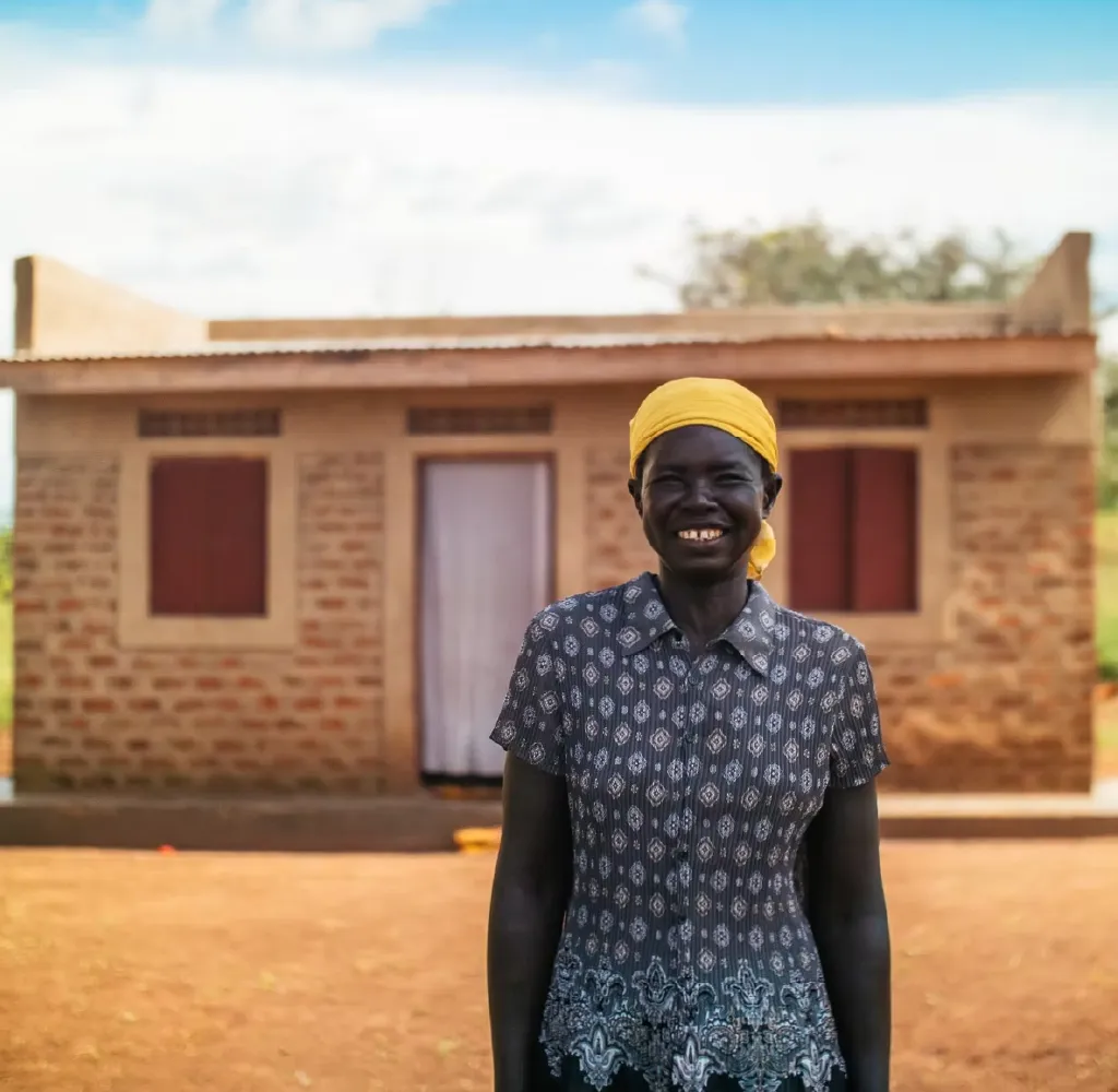 Smiling woman wearing a yellow headscarf and patterned blouse standing in front of a brick house with closed windows and a doorway.