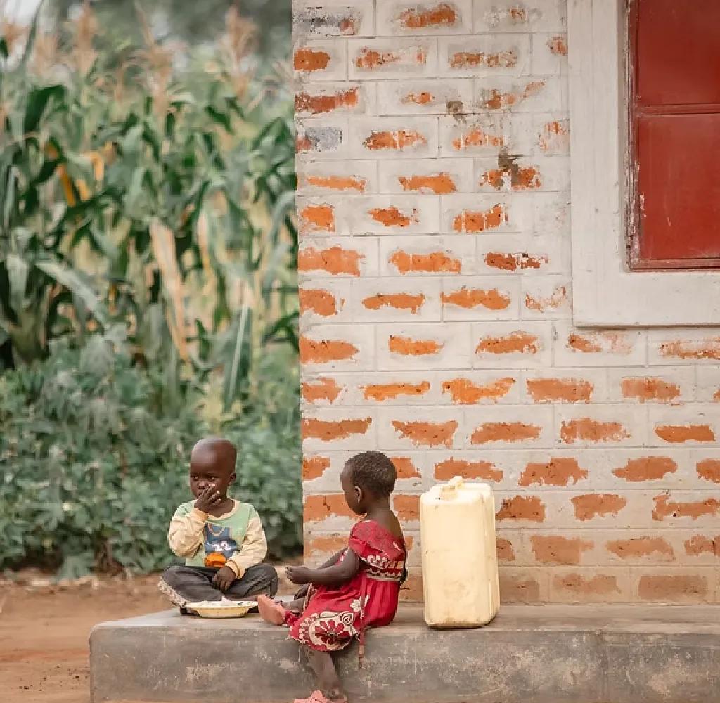 Two young children sitting outside near a brick wall with a yellow water container between them; one child is eating while the other looks on.