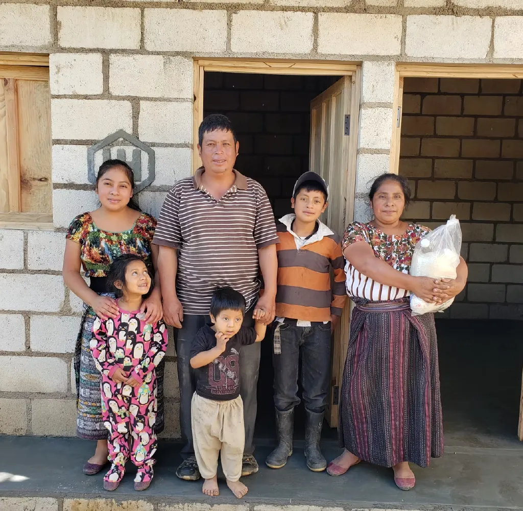 Group of six people, including three children and three adults, standing in front of a stone house; one woman is holding a plastic bag with groceries.