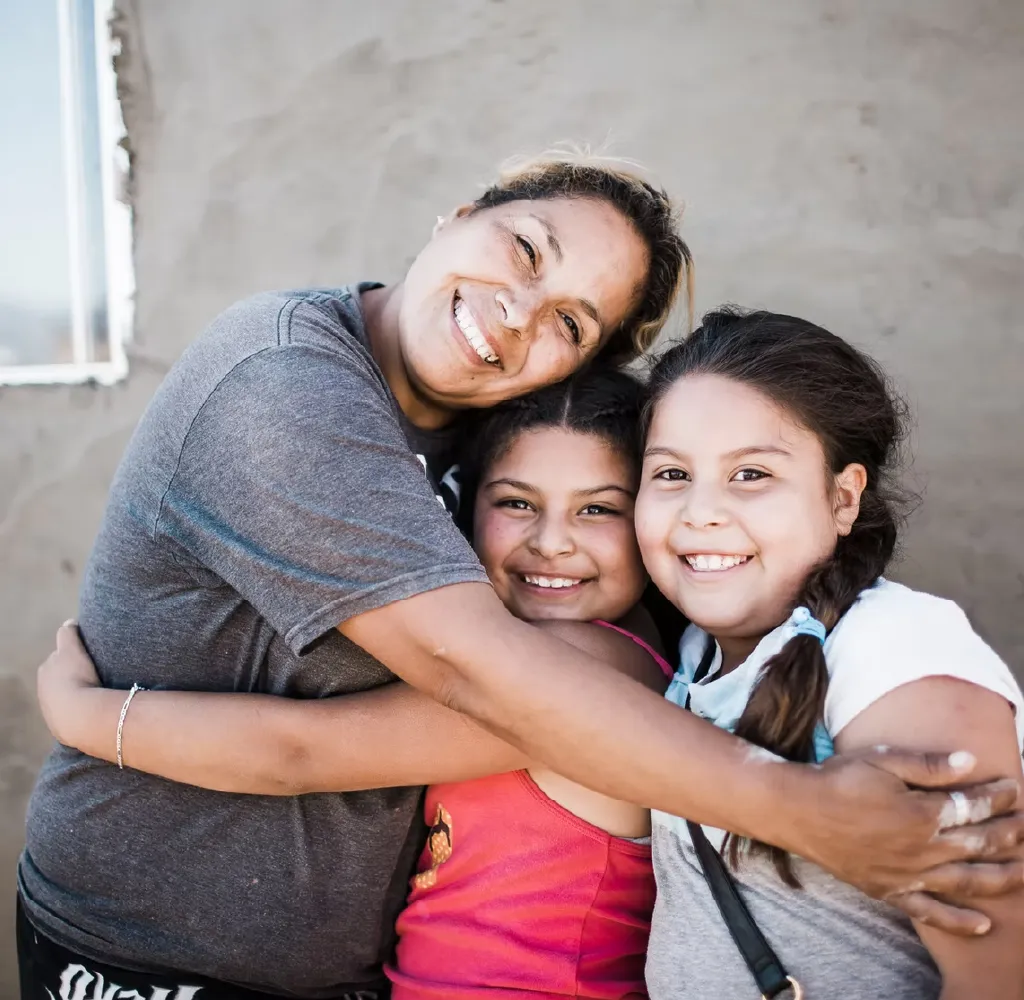 Smiling woman hugging two happy young girls against a plain wall background.