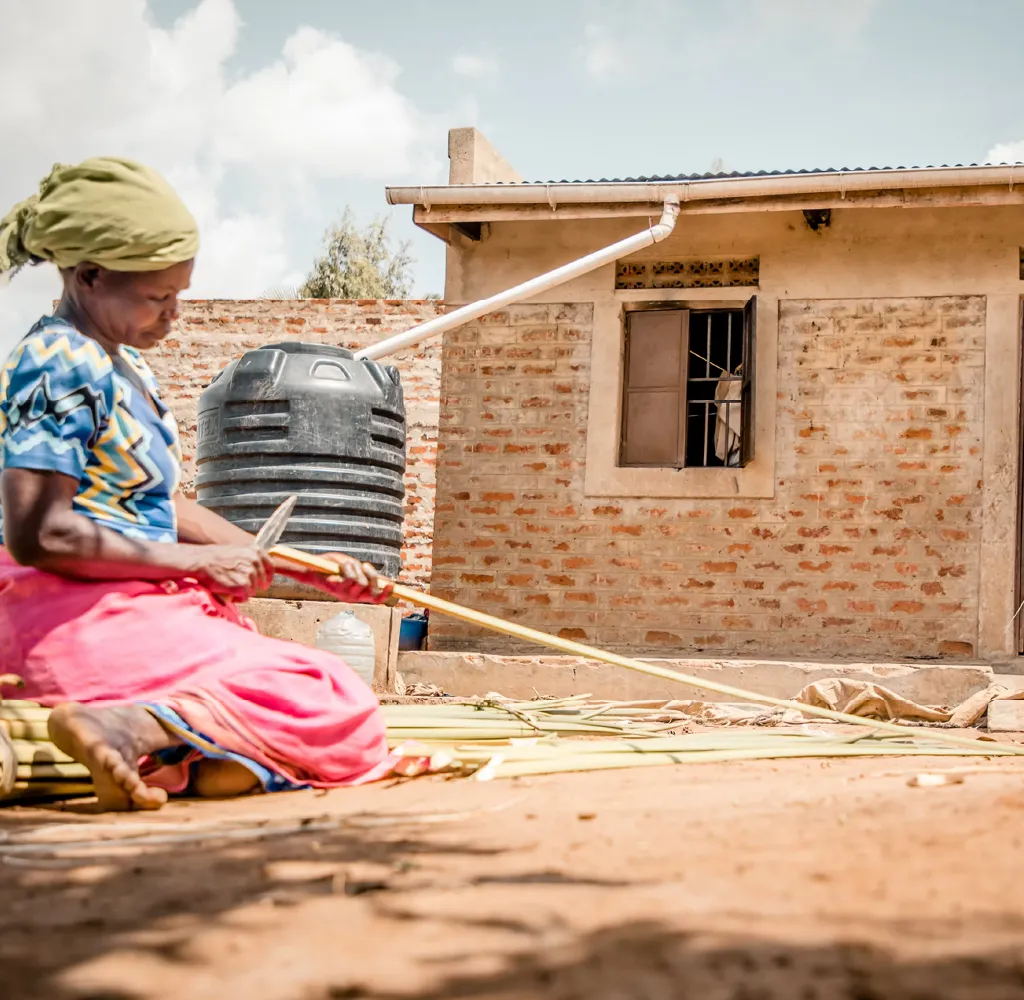 Woman in colorful clothing sitting on the ground peeling or preparing long sticks outside a brick house with a black water tank.