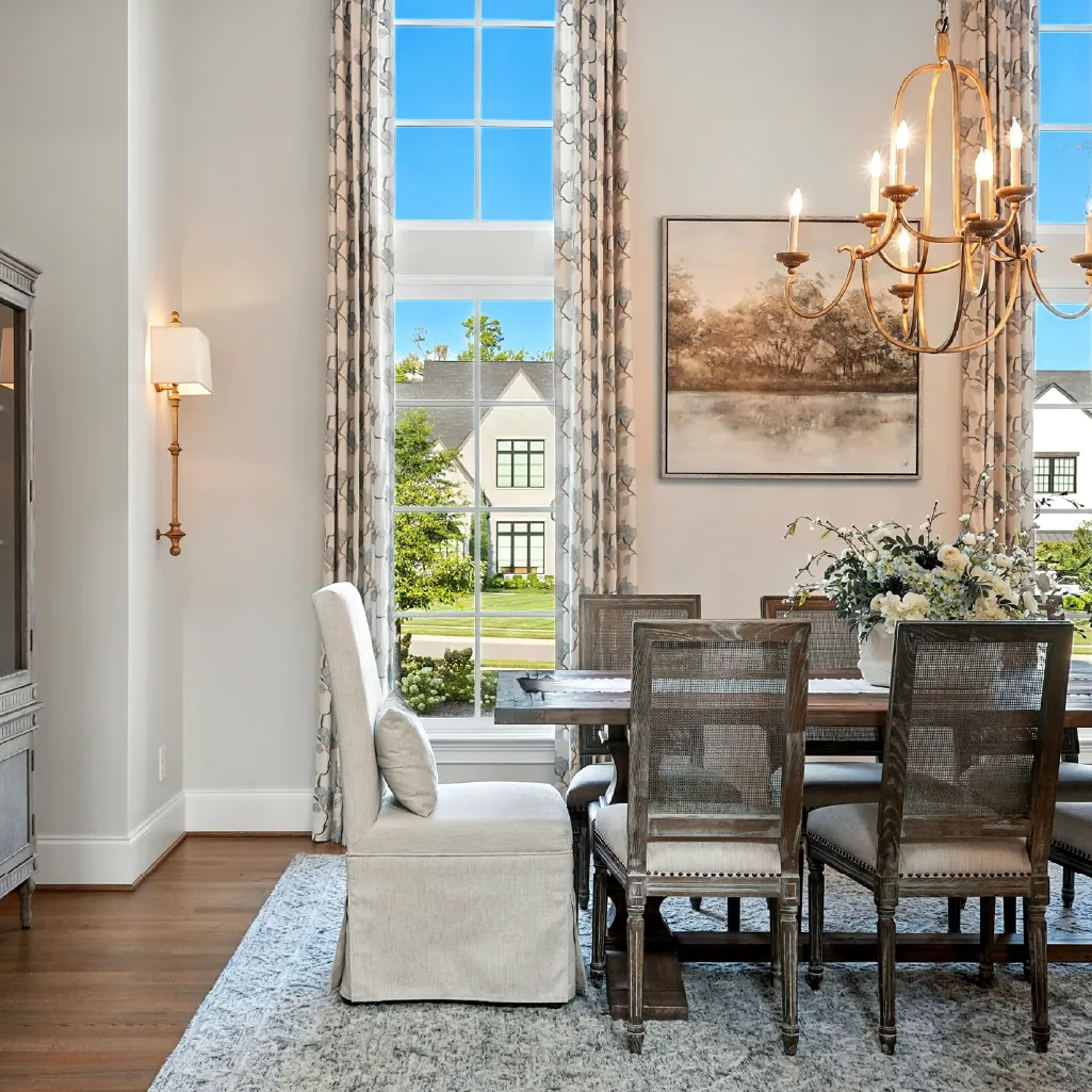 Bright dining room with large windows, floral curtains, wooden table, mixed upholstered and wooden chairs, chandelier, and a wall painting.