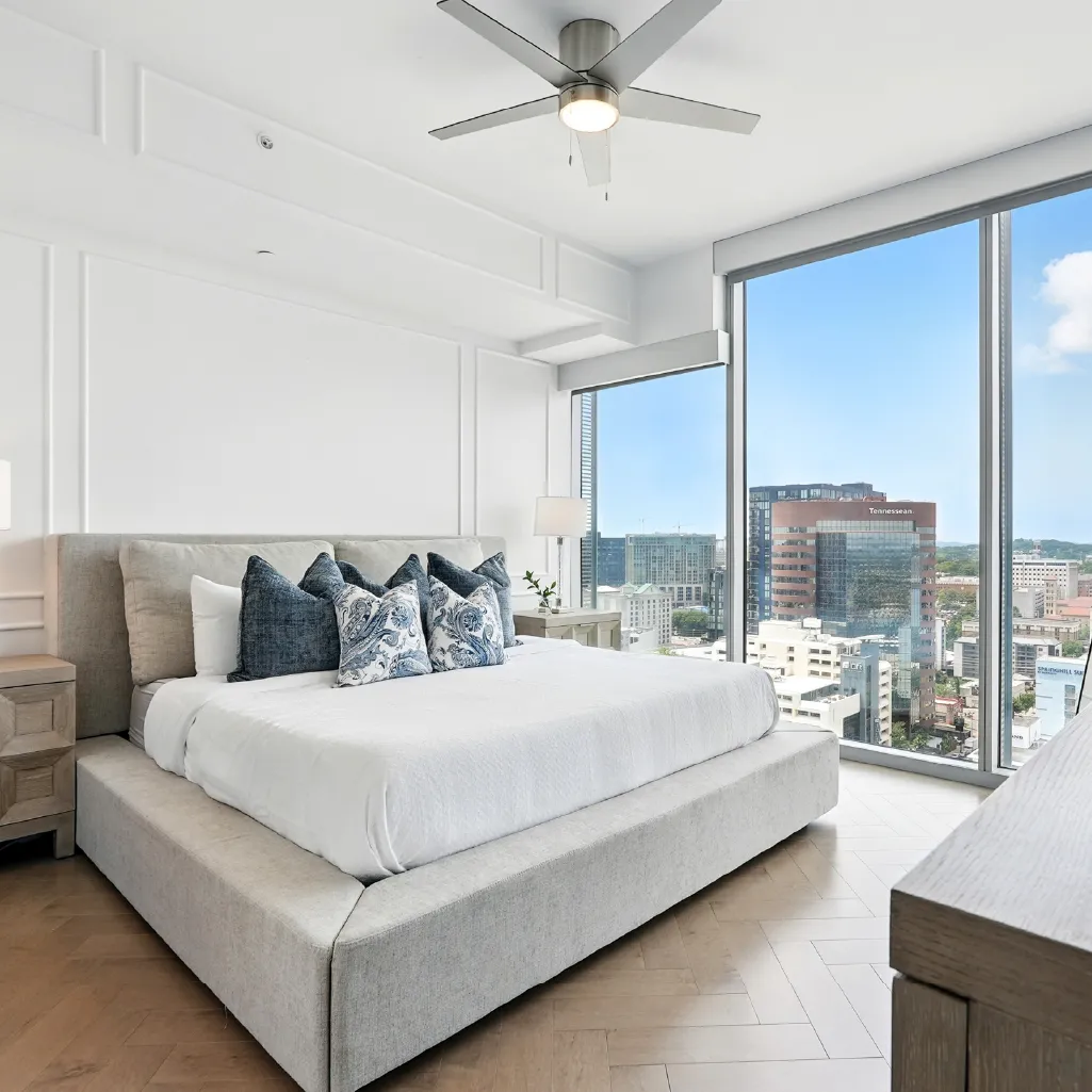 Modern bedroom with large beige bed, blue and patterned pillows, wooden floor, ceiling fan, and floor-to-ceiling windows showing city view.