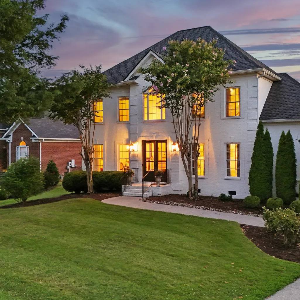 Two-story white brick house with lit windows and front entrance flanked by trees and a green lawn at dusk.