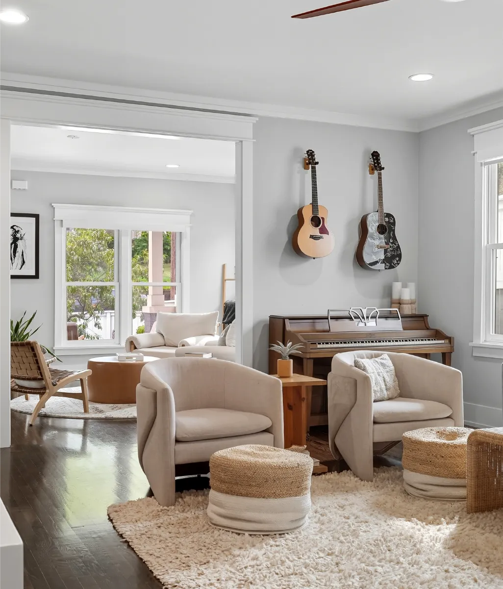 Cozy living room with light armchairs, woven ottomans, a wooden piano under two hanging guitars, and soft rug on dark hardwood floor.
