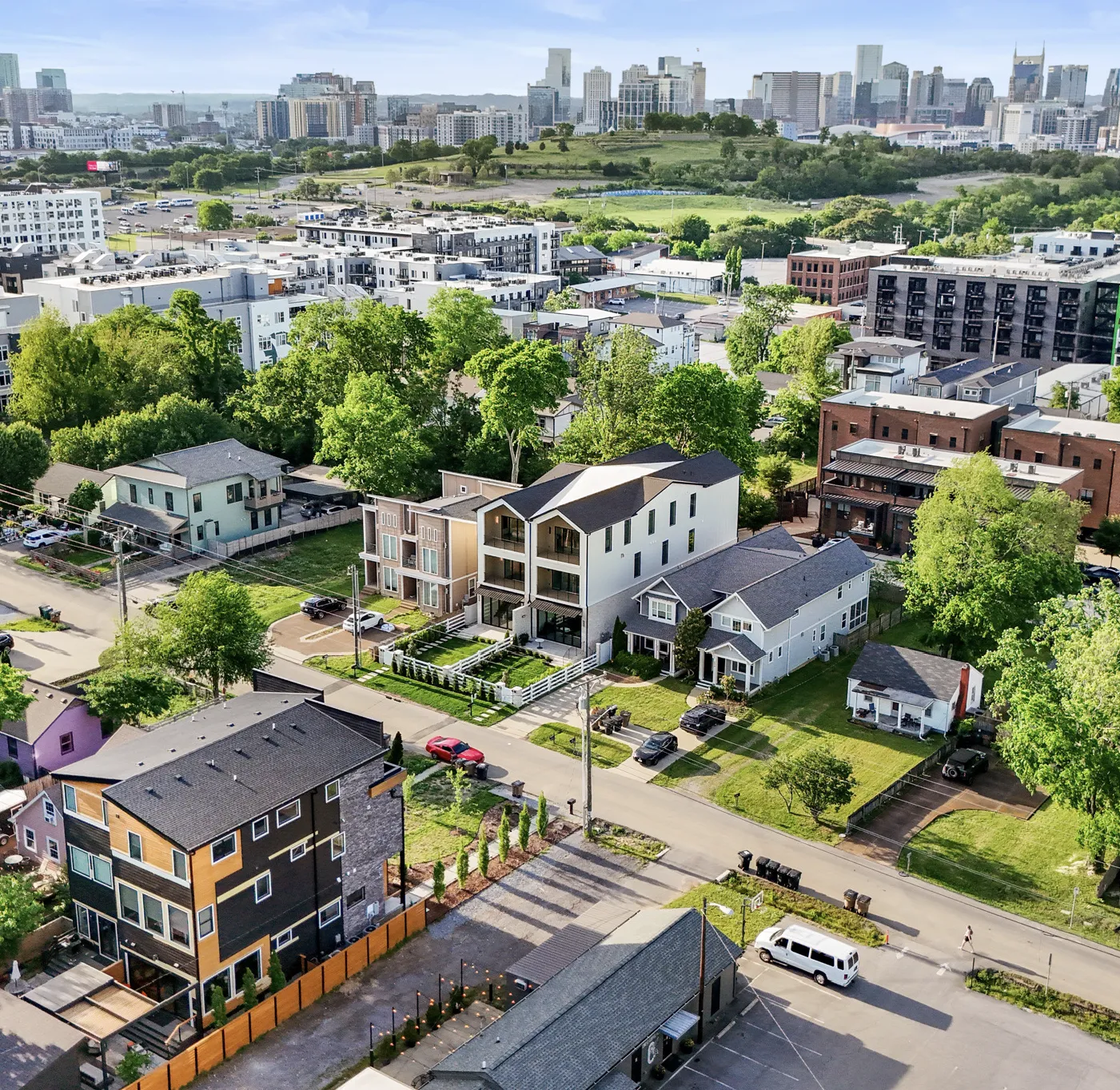 Aerial view of a residential neighborhood with houses, trees, parked cars, and a city skyline in the background under a clear sky.