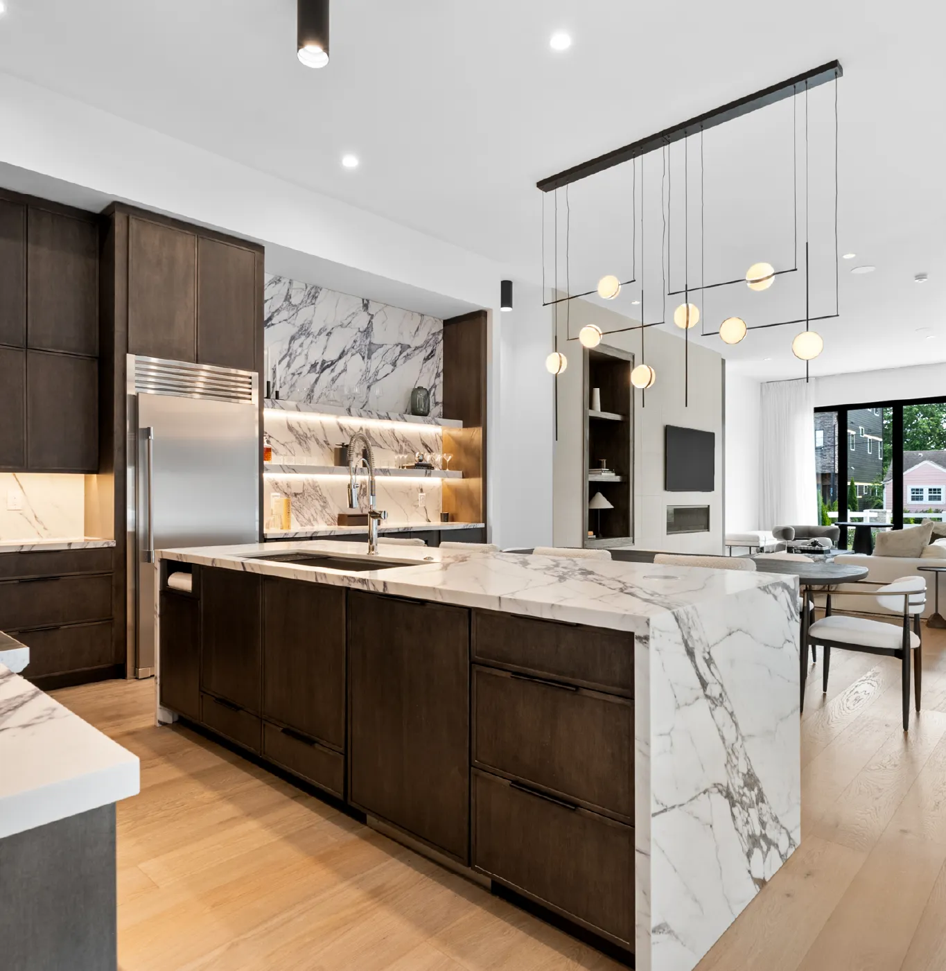 Modern kitchen with dark wood cabinets, white marble countertops and backsplash, an island with sink, and contemporary pendant lights, adjoining a bright living area with large windows.