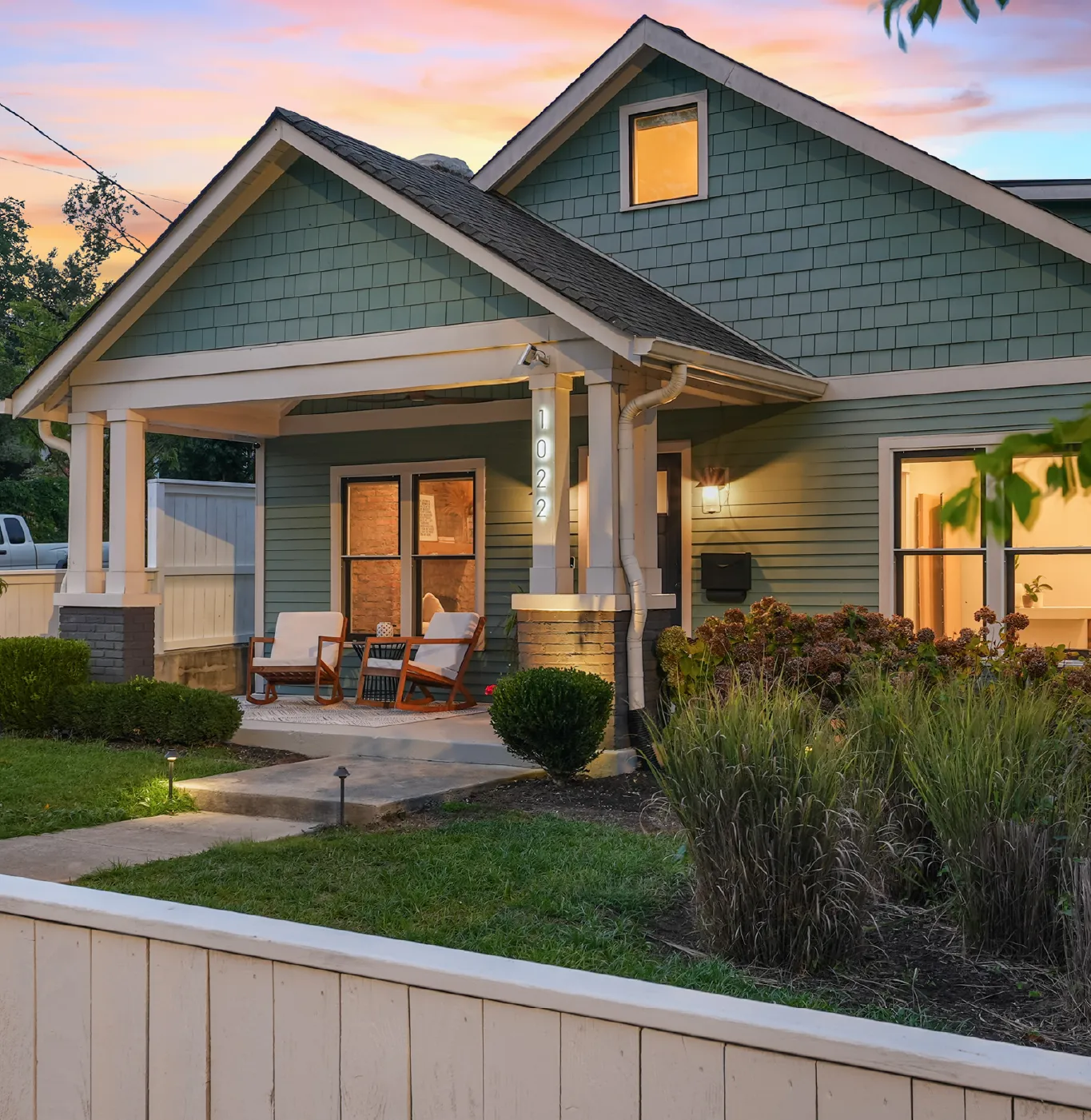 Cozy green house with a front porch featuring two cushioned wooden chairs, lit warmly at twilight.