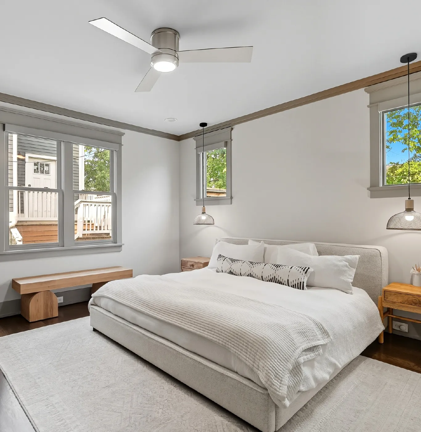 Modern bedroom with a large upholstered bed dressed in white linens, wooden nightstands, pendant lights, ceiling fan, and large windows with a view of greenery and a neighboring house.