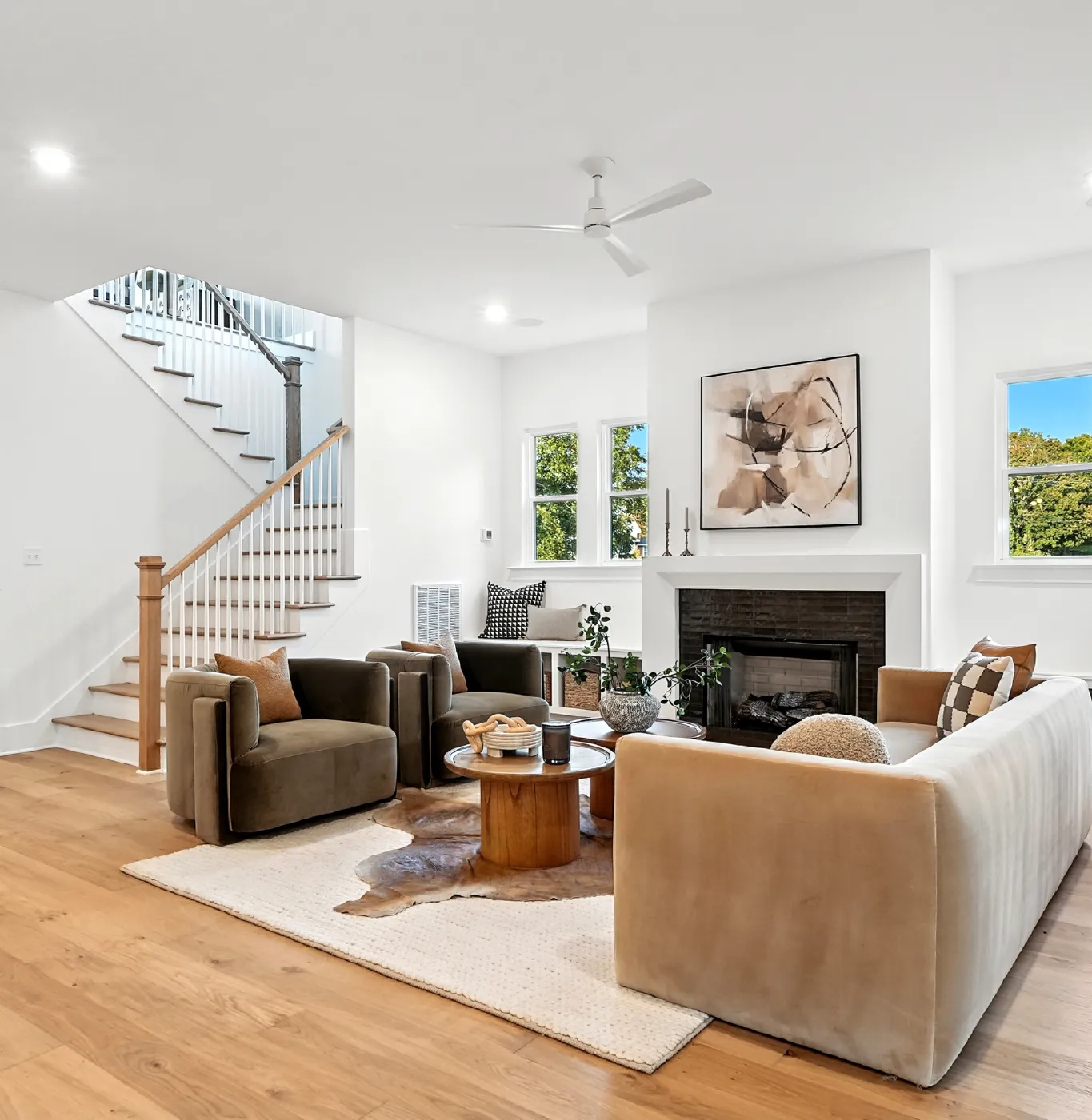 Modern living room with beige sofa, two brown armchairs, round wooden coffee tables, white walls, a fireplace with abstract art above, and a staircase with wooden handrail.