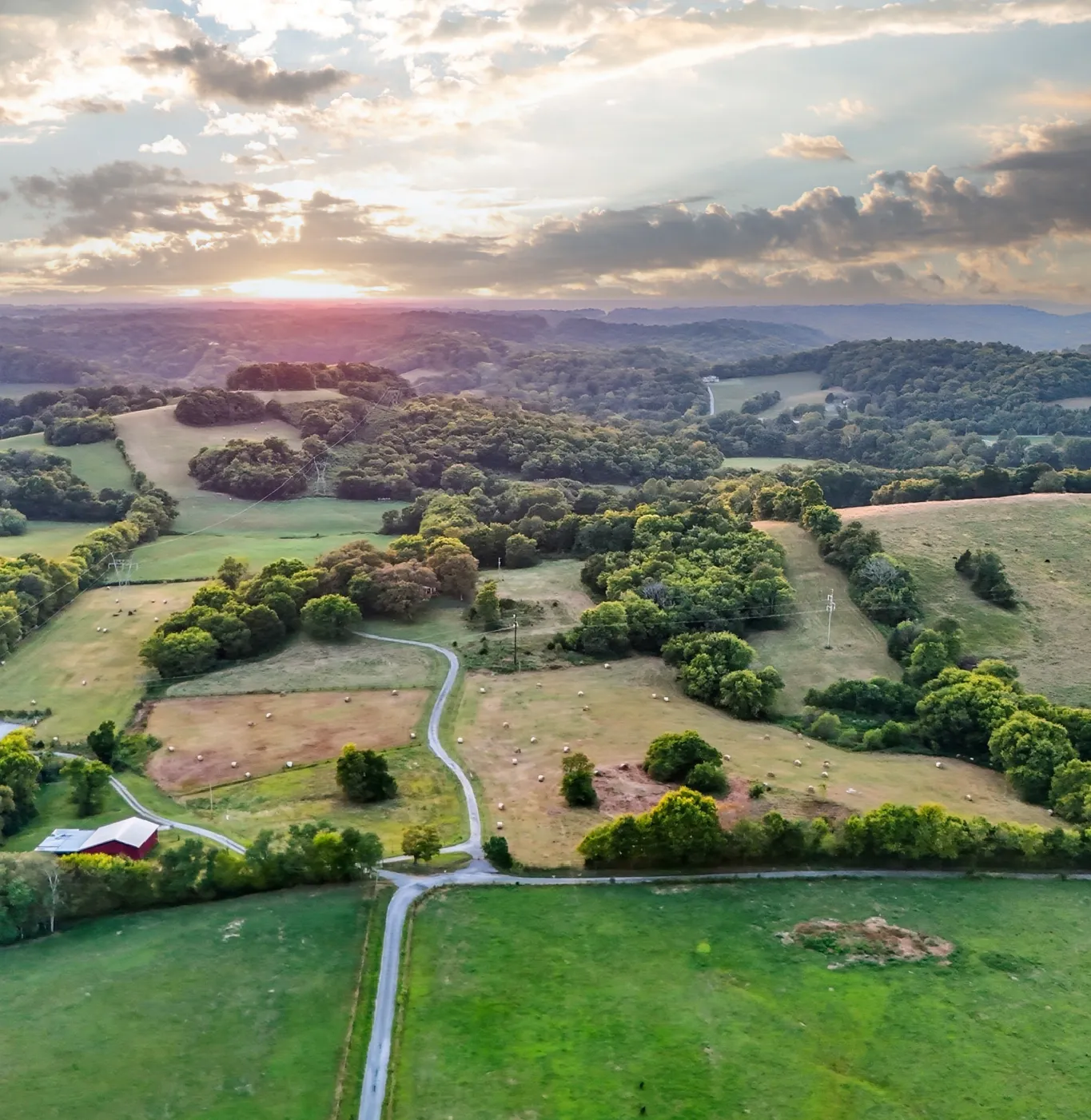 Aerial view of rolling green hills and farmland with a winding road, scattered trees, and a sunset on the horizon.