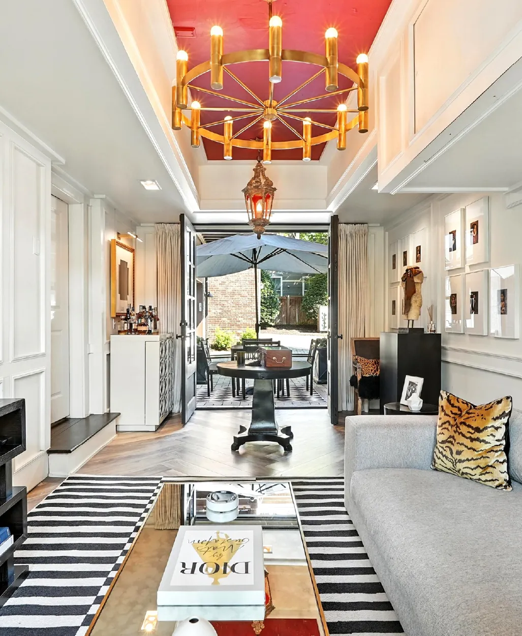 Bright living room with grey sofa, tiger-print pillow, striped rug, gold chandelier, and open doors leading to an outdoor patio with umbrella and seating.
