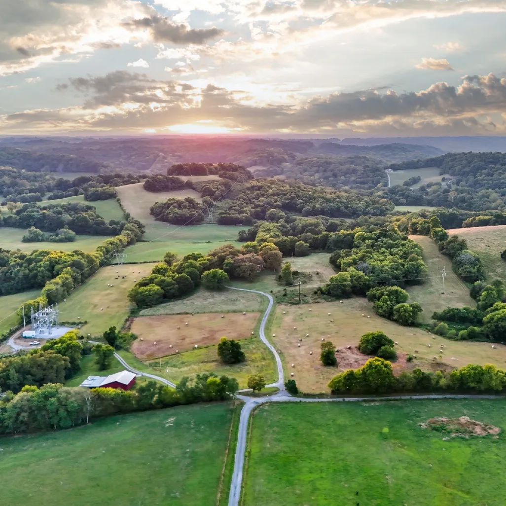 Aerial view of rolling green farmland with scattered trees, hay bales, a red barn, and a winding road under a partly cloudy sky at sunset.