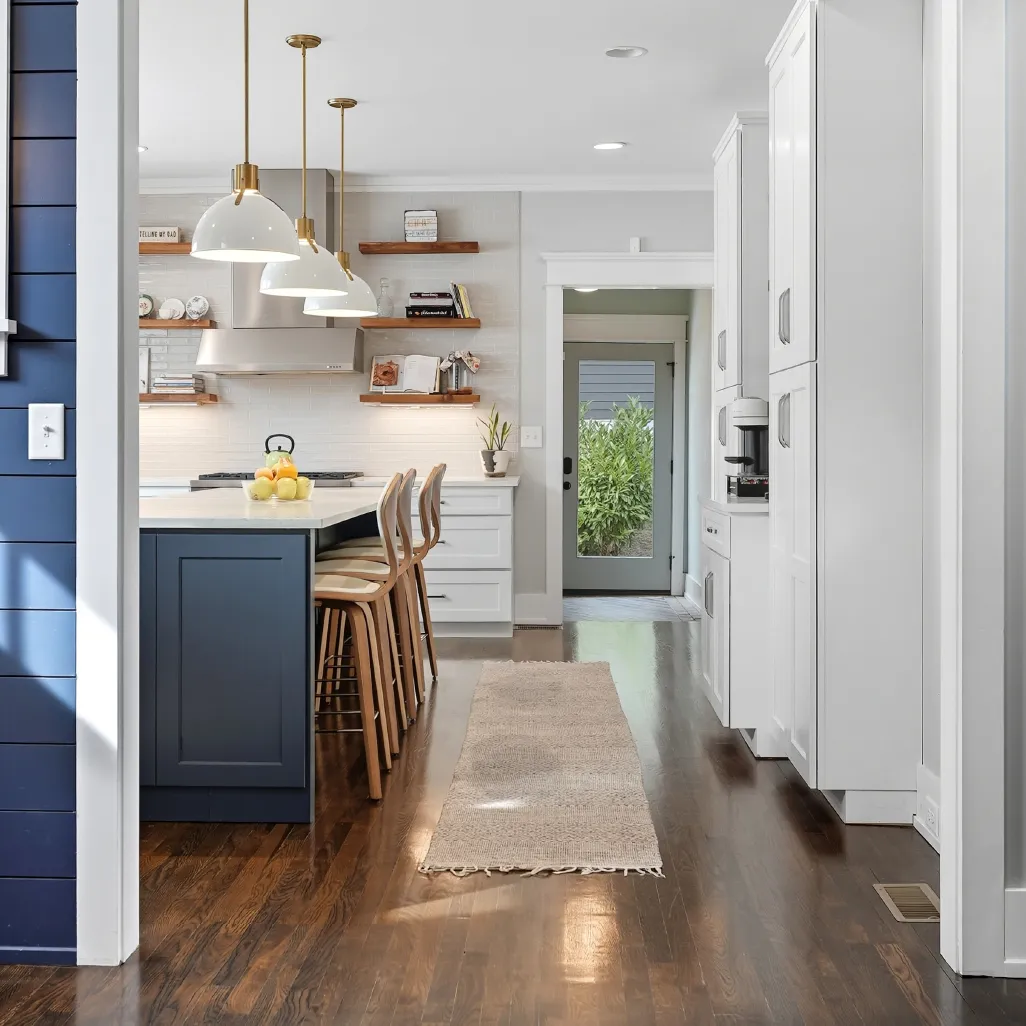 Modern kitchen with dark wood floors, white cabinets, blue island with wooden stools, hanging pendant lights, and glass door leading outside.