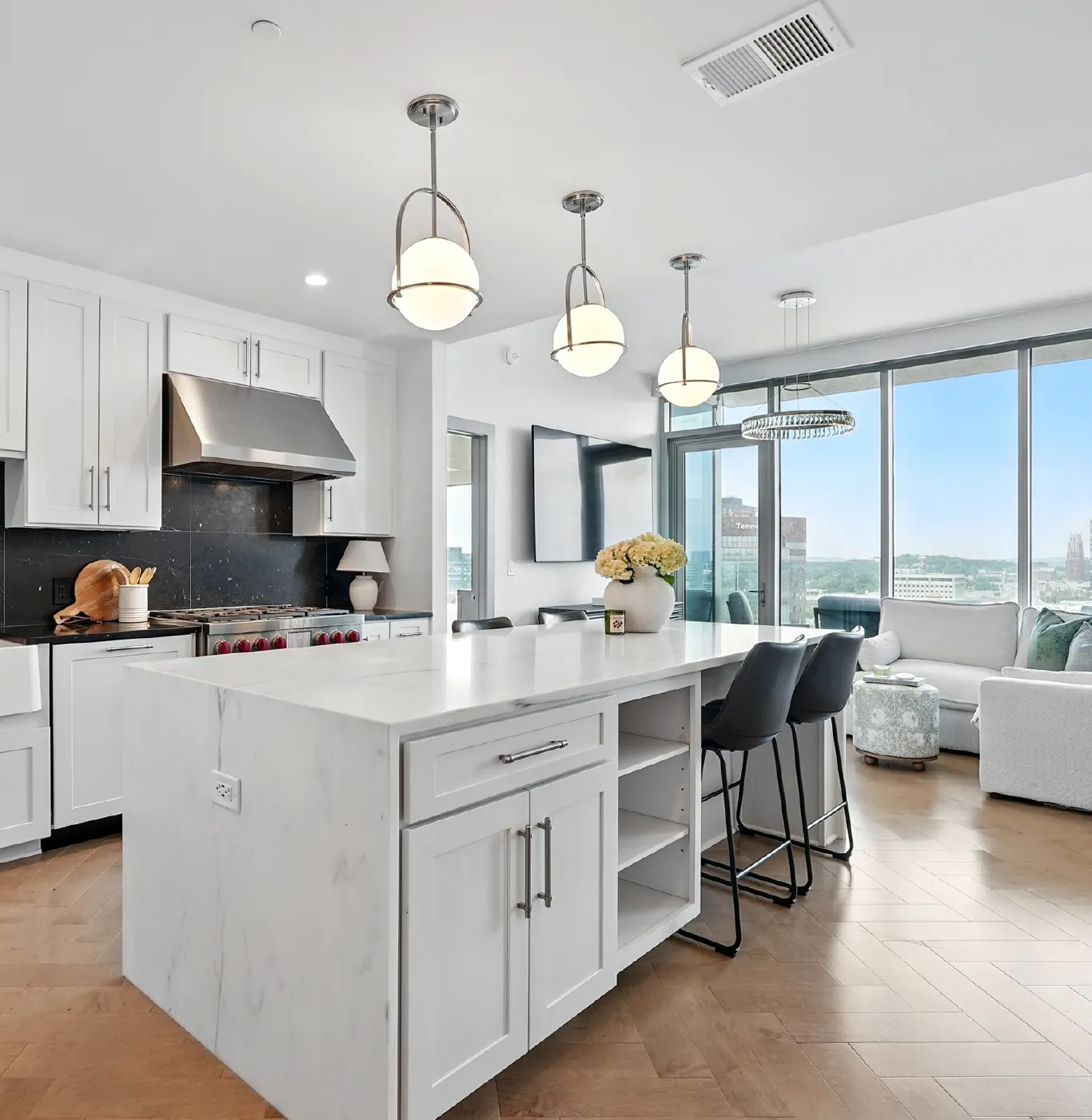 Modern kitchen with white cabinets, marble island with bar stools, pendant lights, and large windows overlooking a city view.