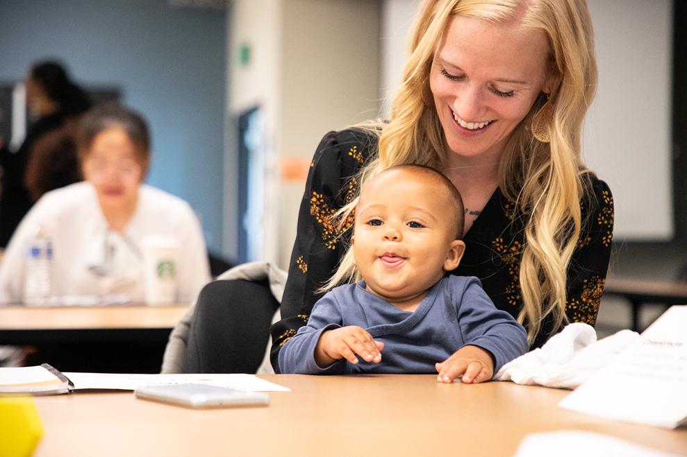 Smiling blonde woman holding a happy baby sitting at a classroom table.